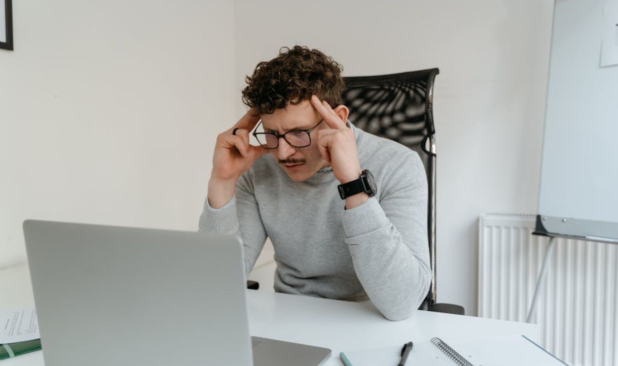 A Man in Gray Sweater Looking at His Laptop with His Hands on His Head