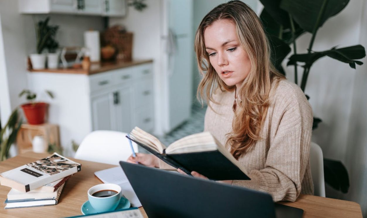 Focused woman checking notebook at table with laptop