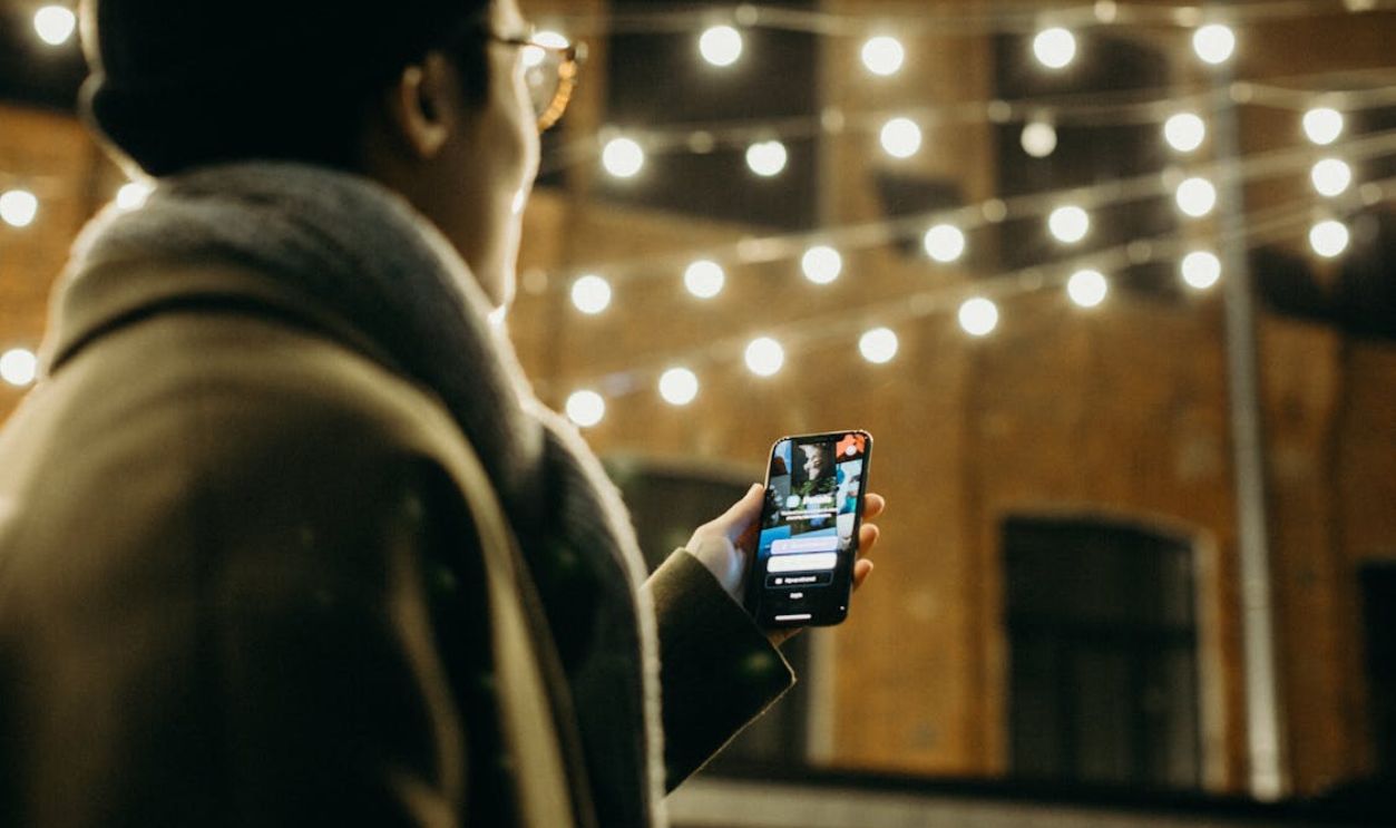 Selective Focus Photography of Man Using Smartphone Beside String Lights