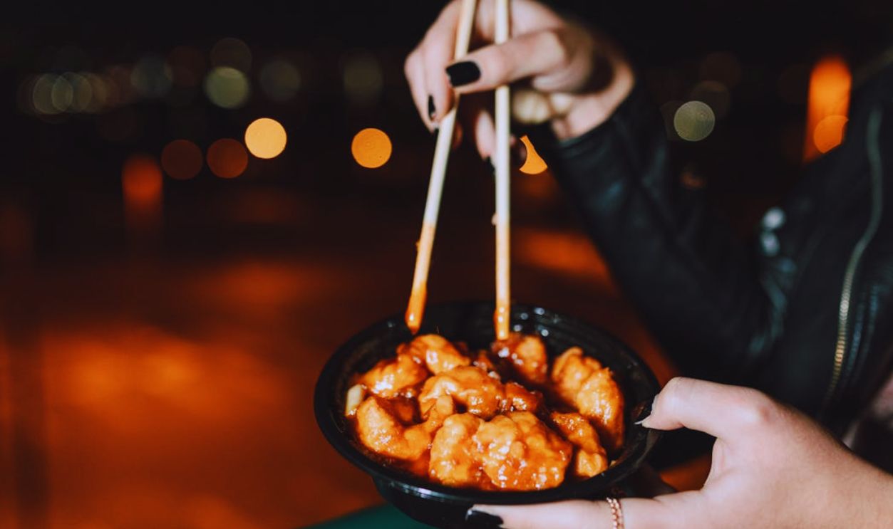 A Person Wearing Black Leather Jacket Eating Meat with Orange Sauce Using Chopsticks