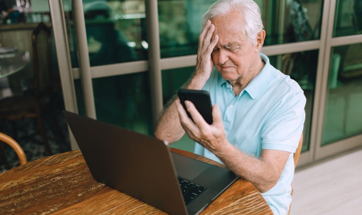 Elderly Man Sitting with Laptop and Holding Hand on Forehead