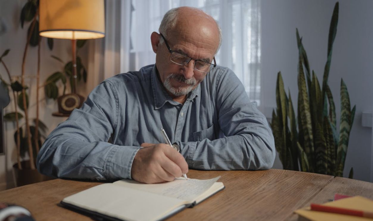 Man in Gray Dress Shirt Wearing Black Framed Eyeglasses Writing on White Paper