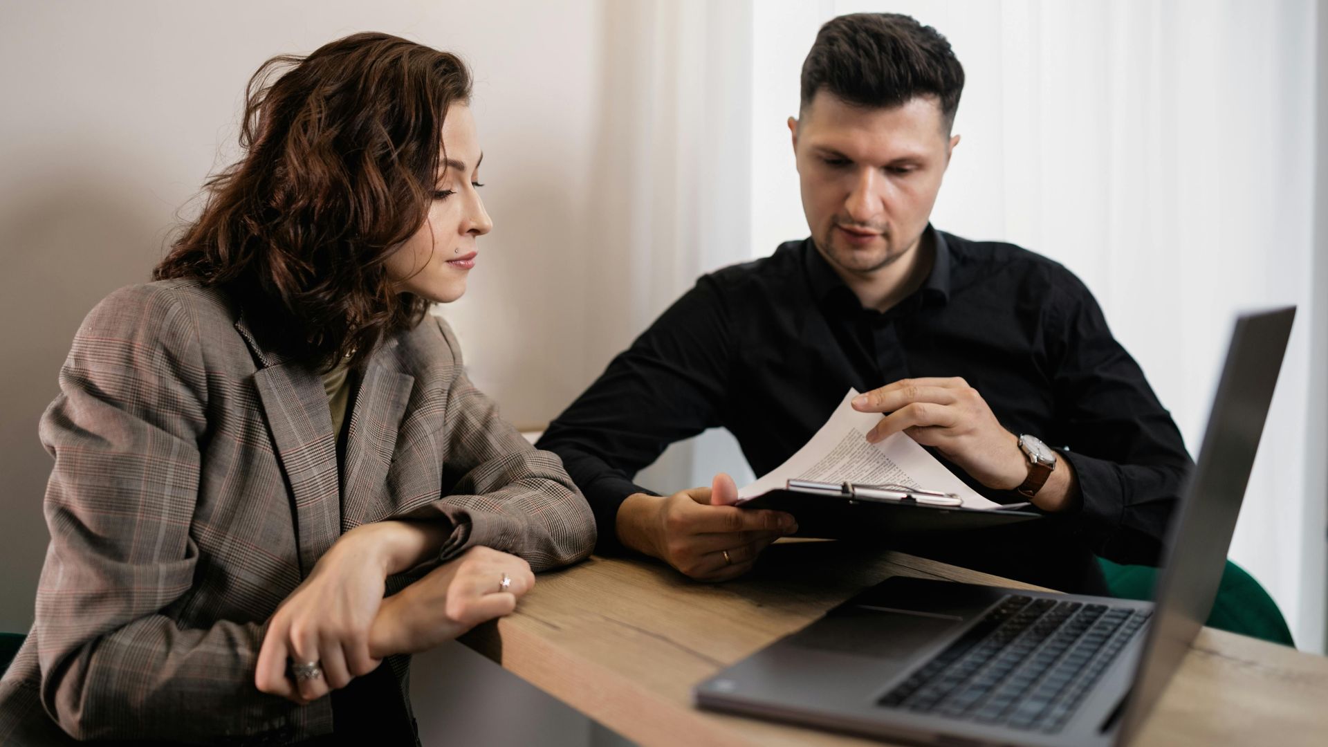 Two professionals discussing documents at a desk with a laptop, focused on collaboration.
