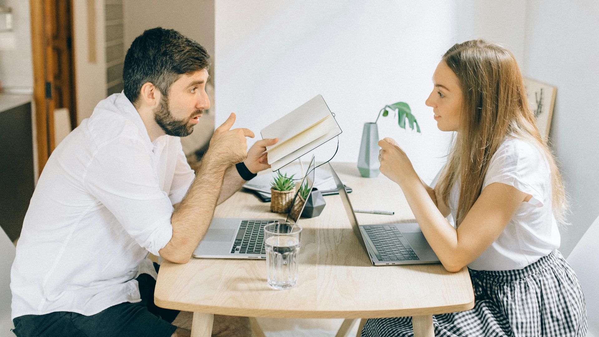A man and woman working from home on laptops, engaged in discussion at a wooden table.