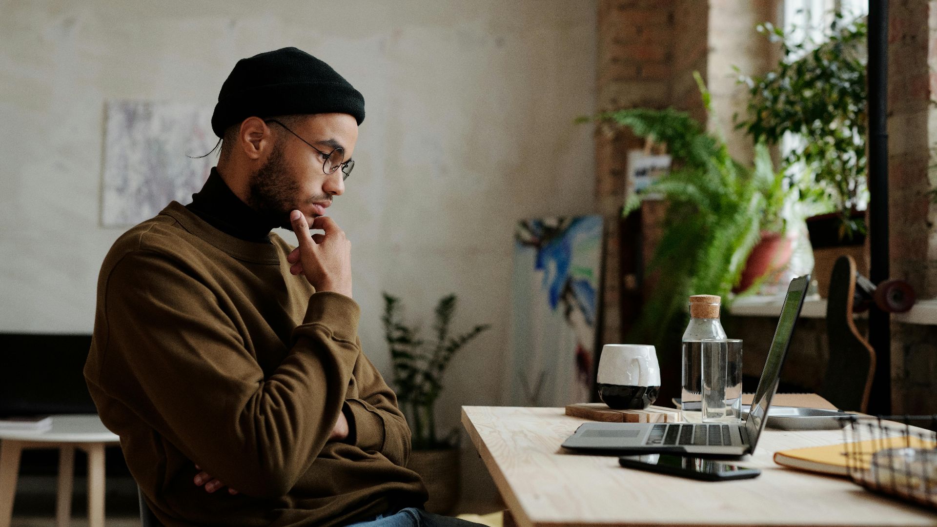 A man in casual attire working thoughtfully on a laptop in a cozy home setting with plants.