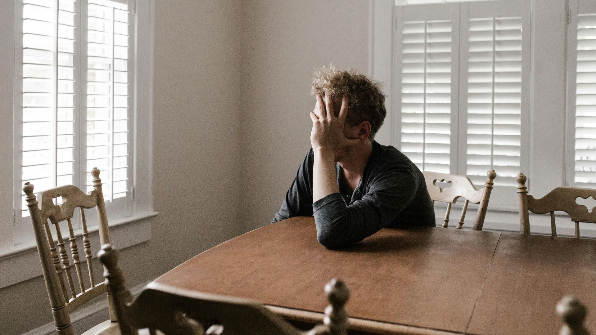 A man sits alone at a table in a bright room, displaying deep contemplation.