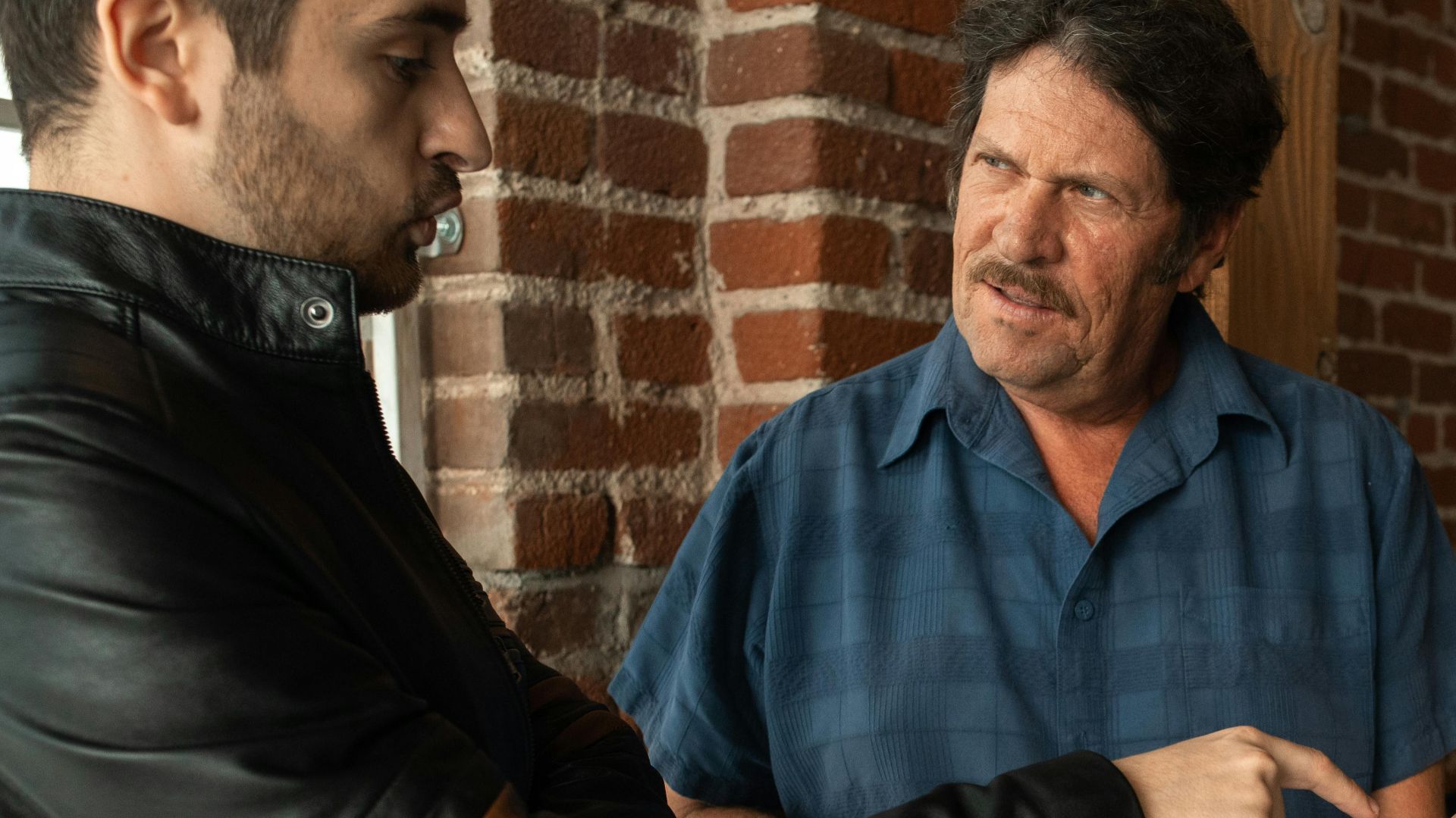 Two men having a conversation indoors, using a smartphone amidst a brick wall setting.