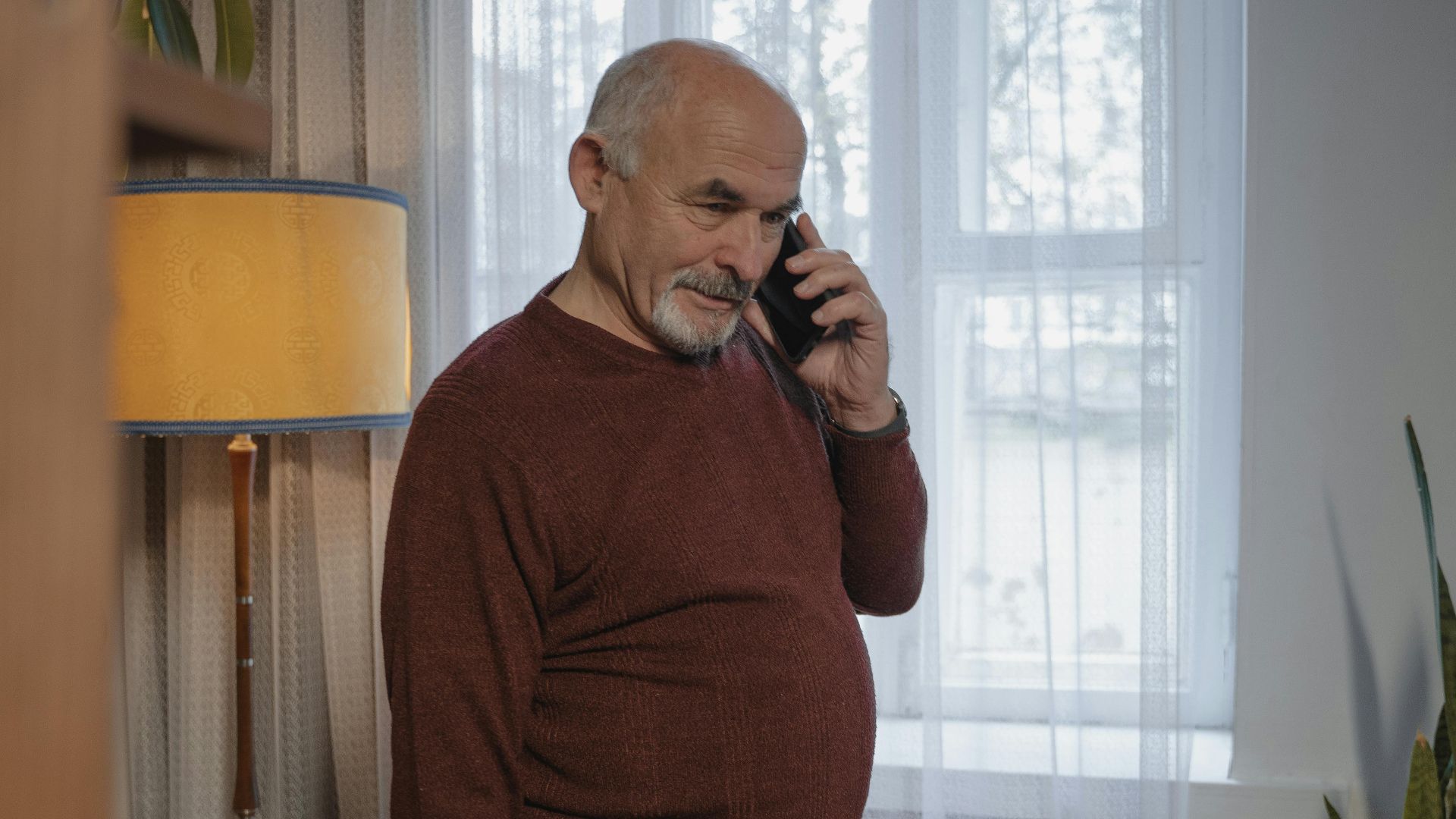 Senior man in brown sweater conversing on phone inside a warmly lit room.