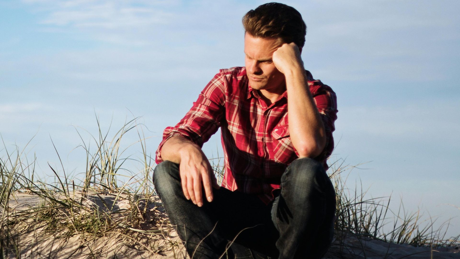Man in a red plaid shirt sitting thoughtfully on a sandy dune in Australia, surrounded by grass.