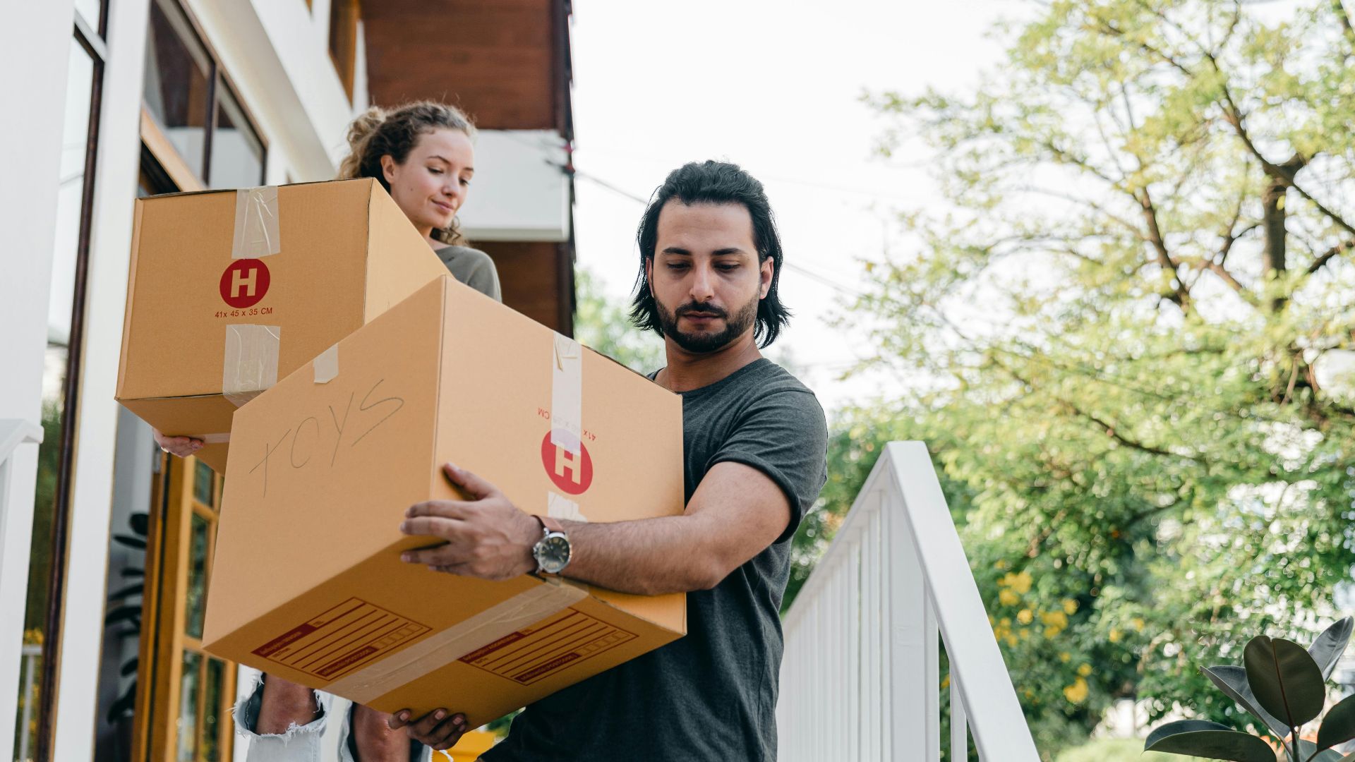 Young multiracial boyfriend and girlfriend in casual wear carrying big carton boxes and watching step on porch stairs while moving out of old home on sunny day in summer