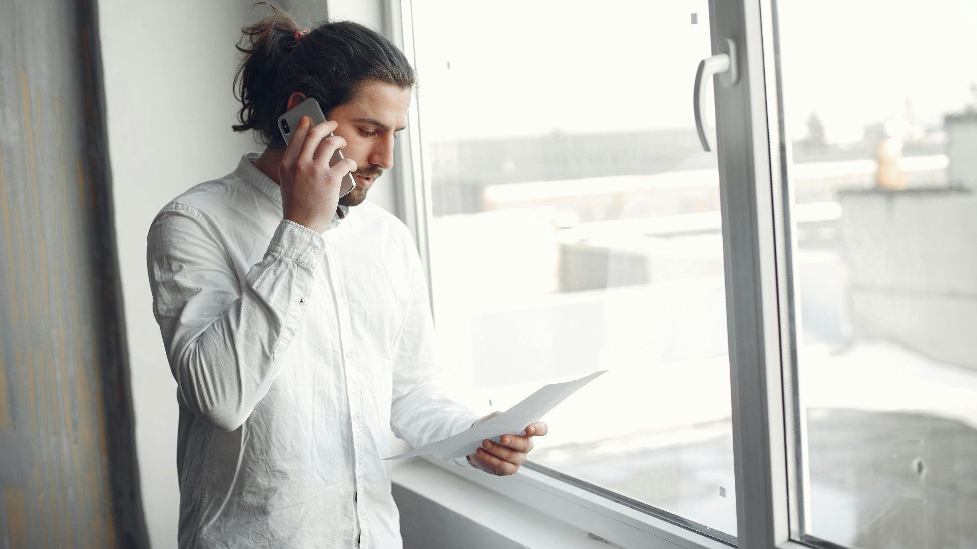 Young man in white shirt, on phone call holding a document, standing by a large window.