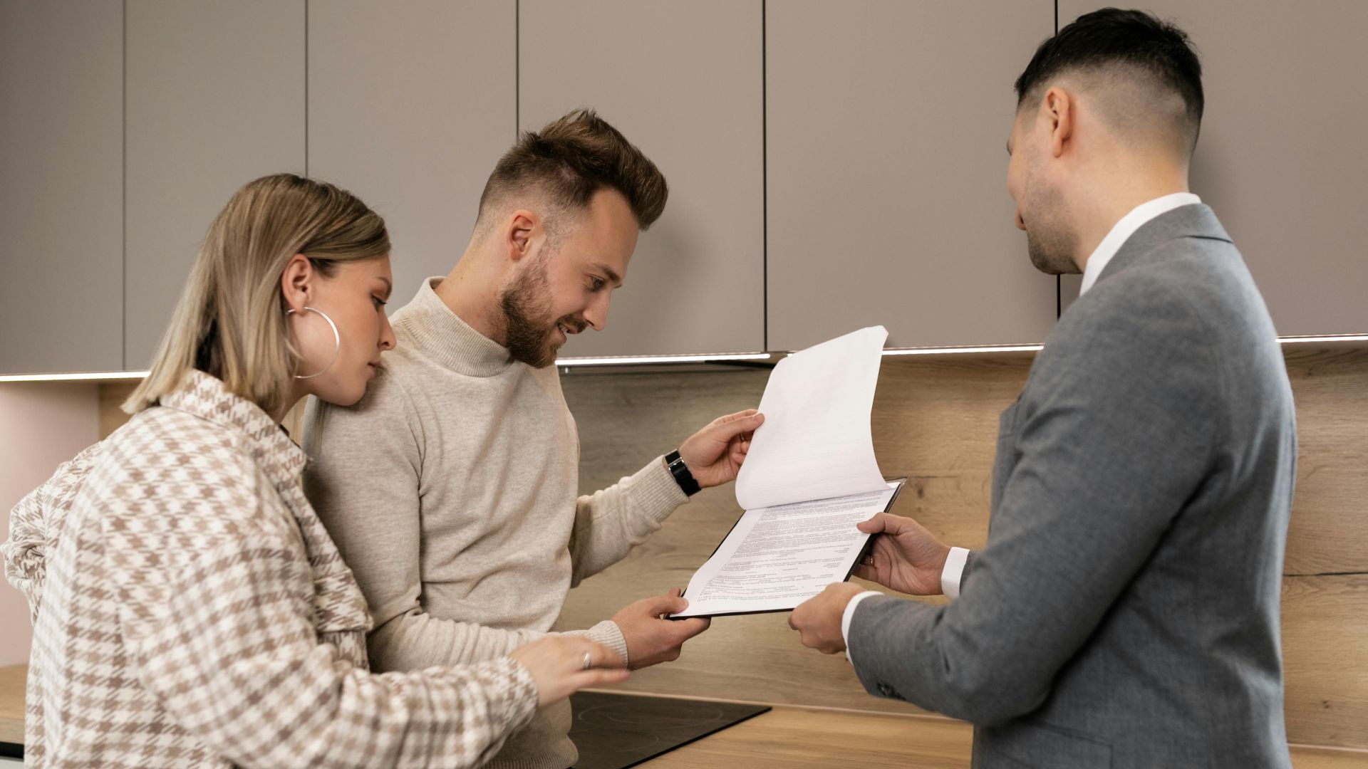 A couple reviews real estate documents with an agent in a modern indoor setting, discussing a potential property purchase.