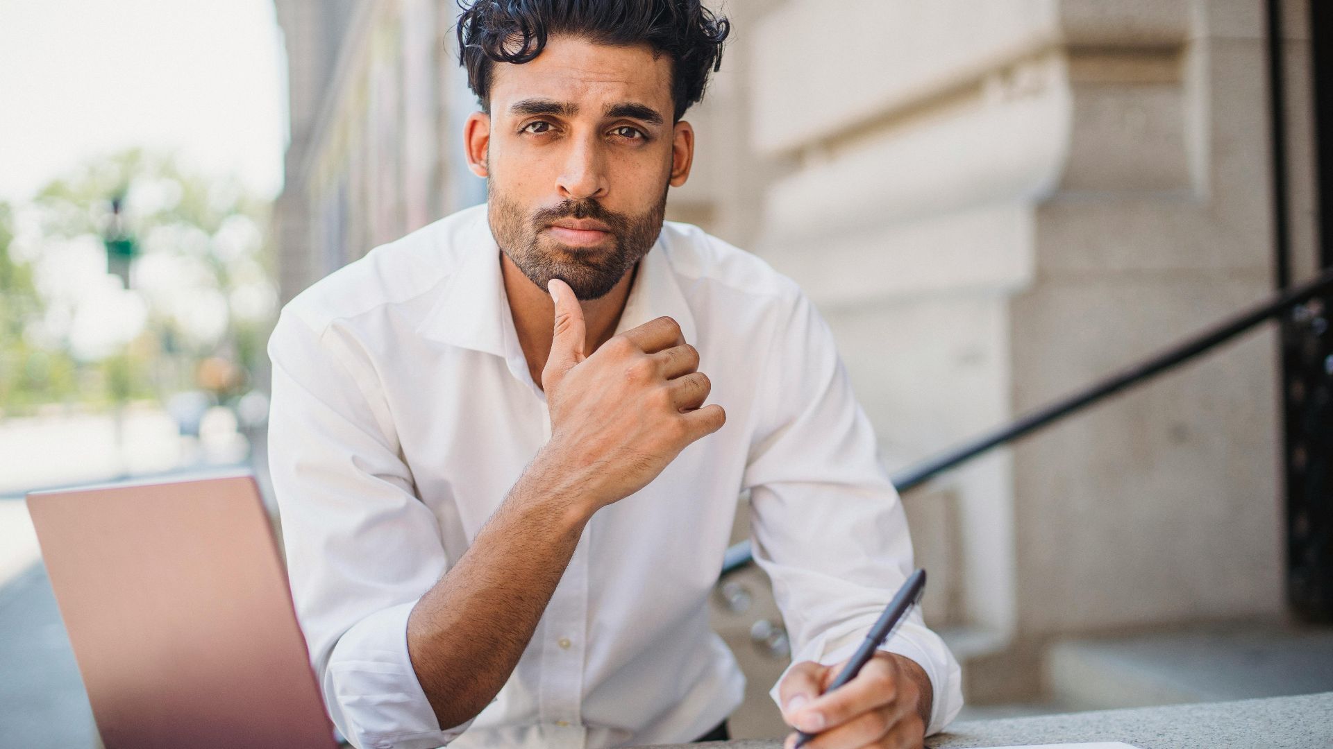 A man in formalwear thinking while writing in a notebook with a laptop outdoors.