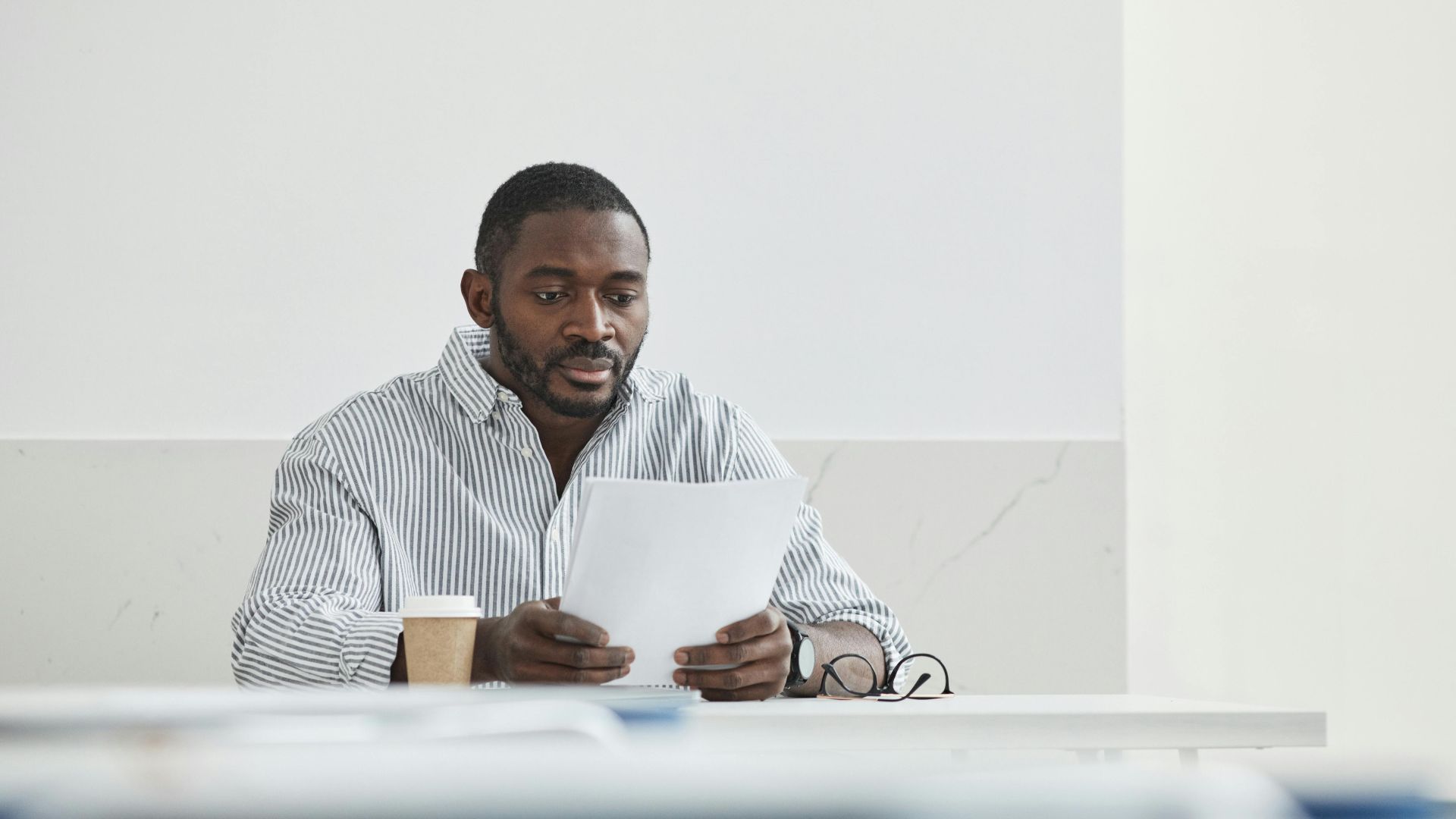 African American man sitting indoors, reading papers with a coffee cup nearby.