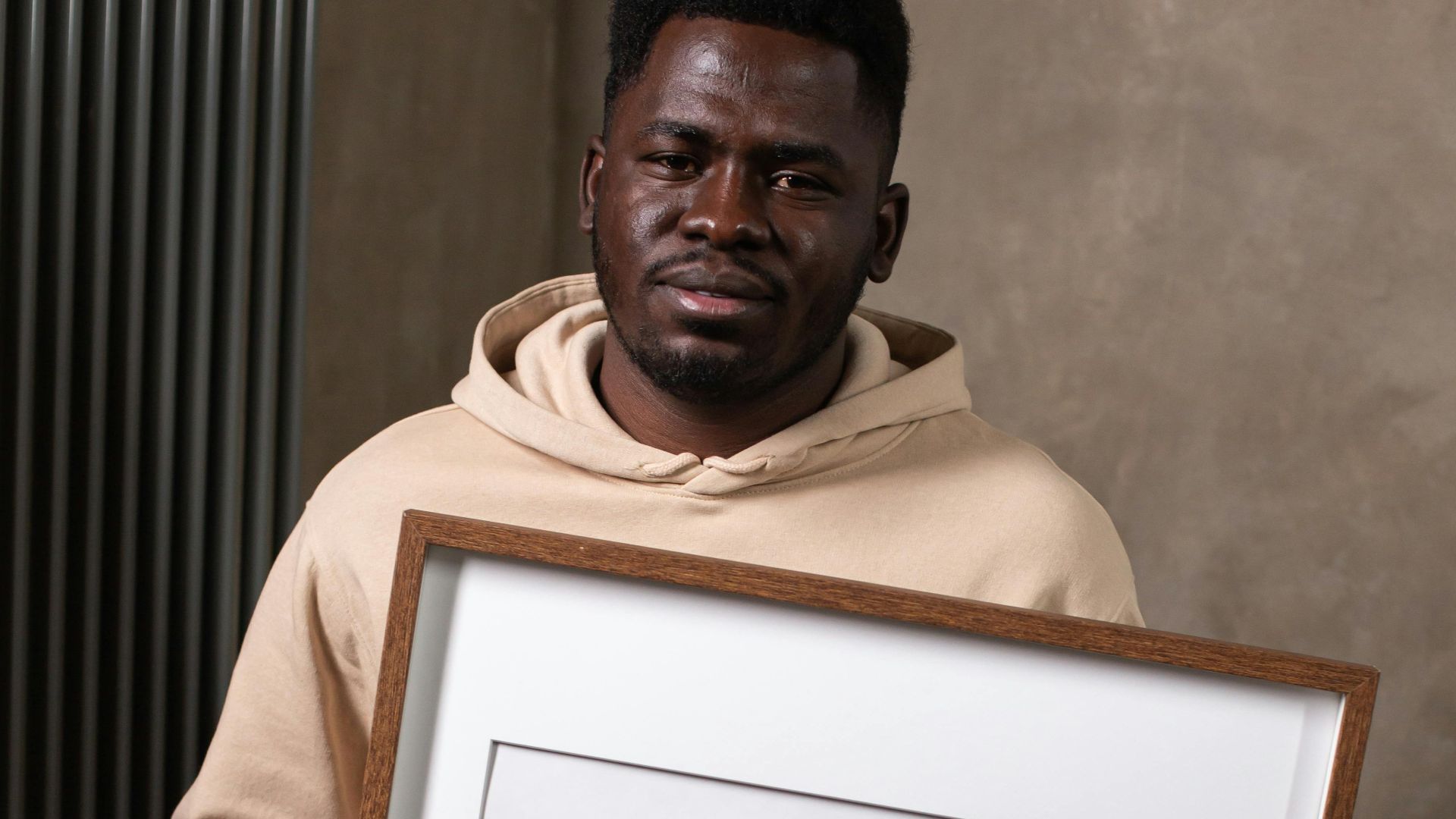 Emotionless African American male in light hoodie holding big empty photo frame in room and looking at camera