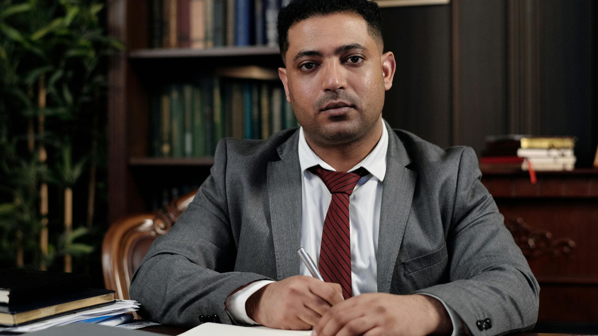 A serious businessman in a suit writing at his desk in a formal office setting.