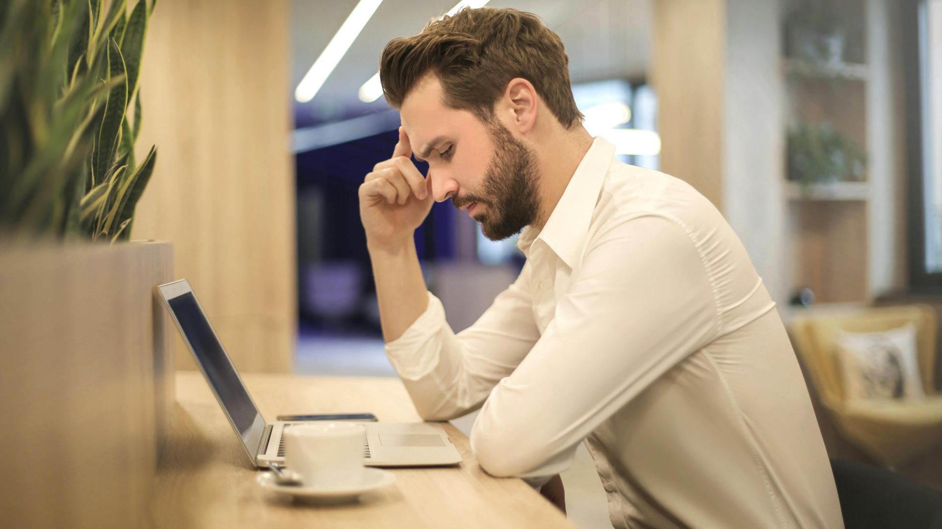 A thoughtful man in a shirt works on his laptop at a modern indoor office space.