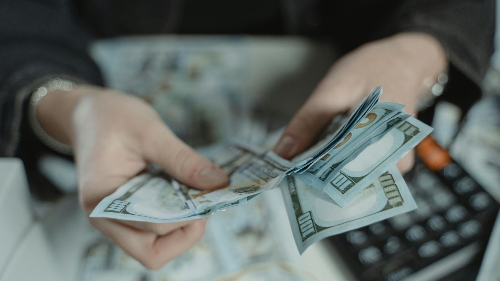 Close-up of hands counting hundred dollar bills with a calculator in the background.