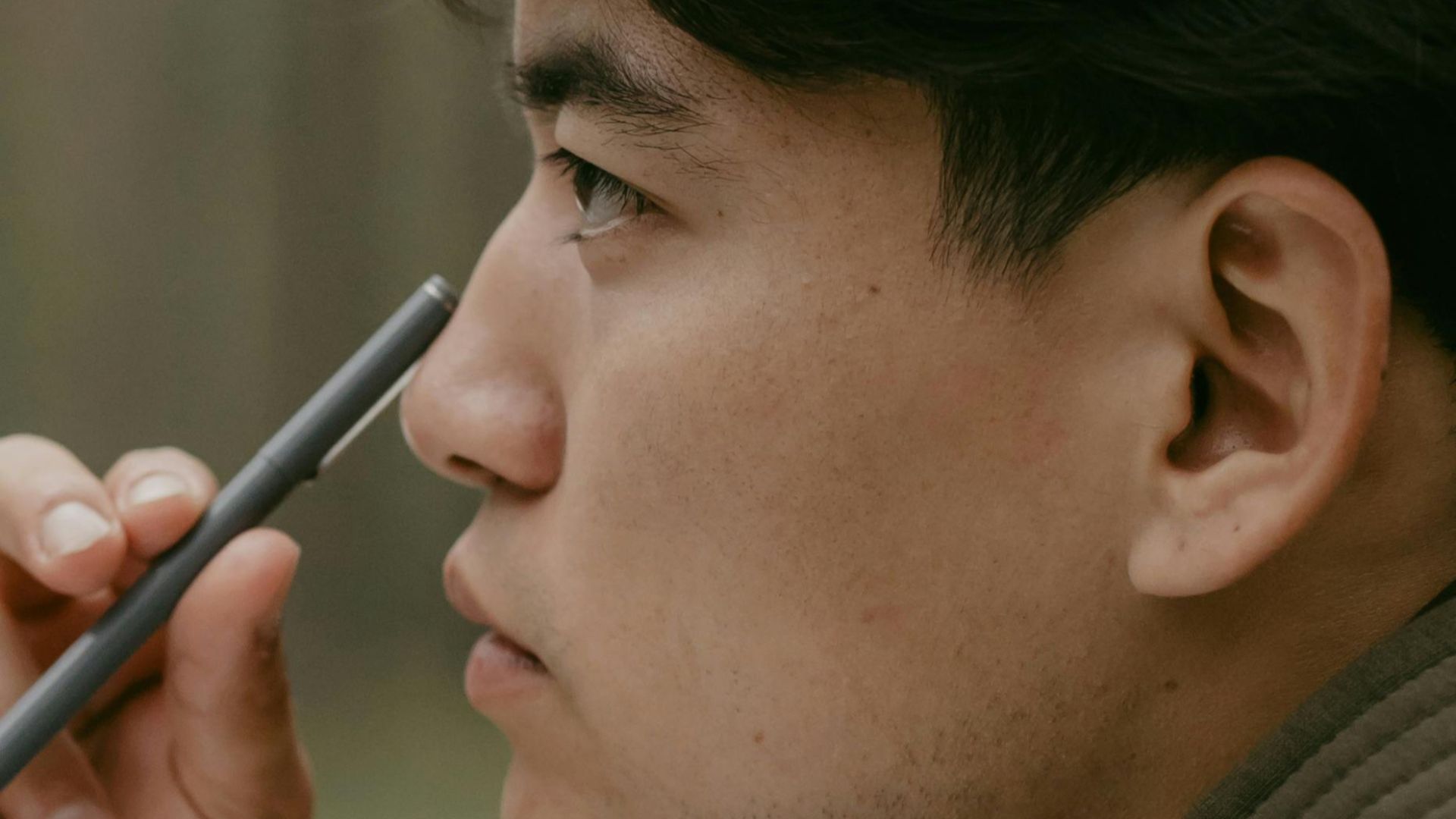 Close-up side view of a man lost in thought, holding pen near face outdoors.