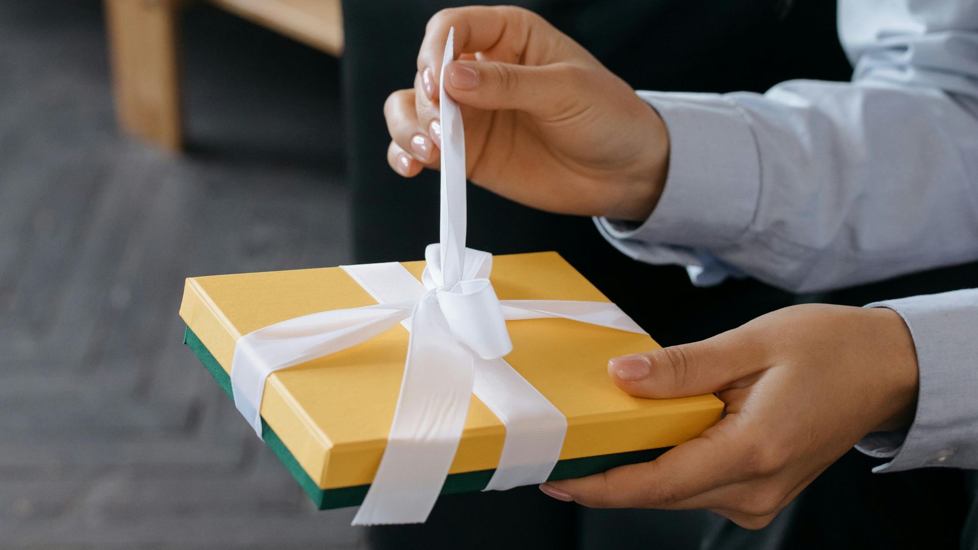 Close-up of hands untying a yellow gift box with a white ribbon indoors.
