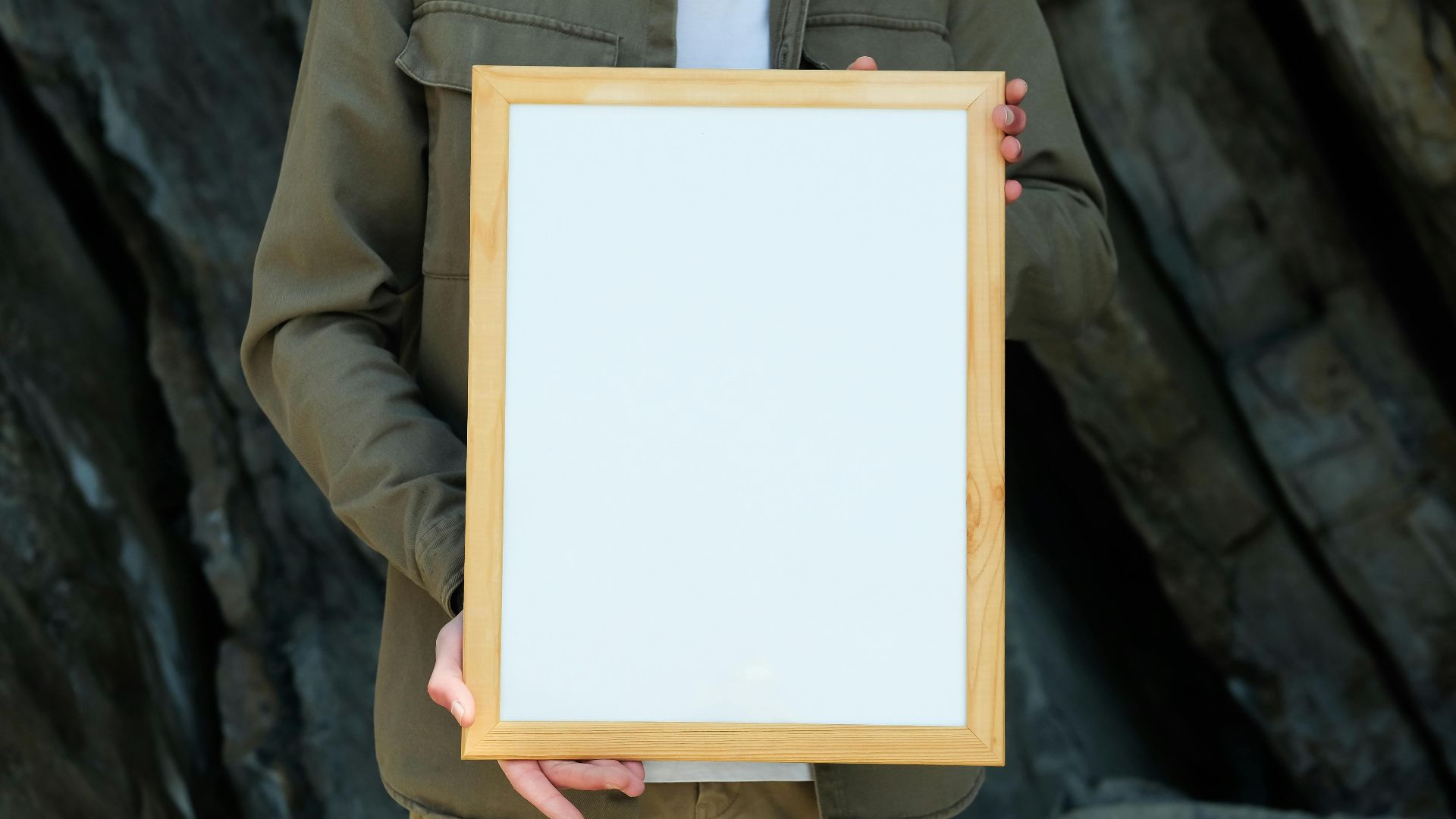 A person holding a blank wooden frame against a rocky outdoor background, ideal for mockups.