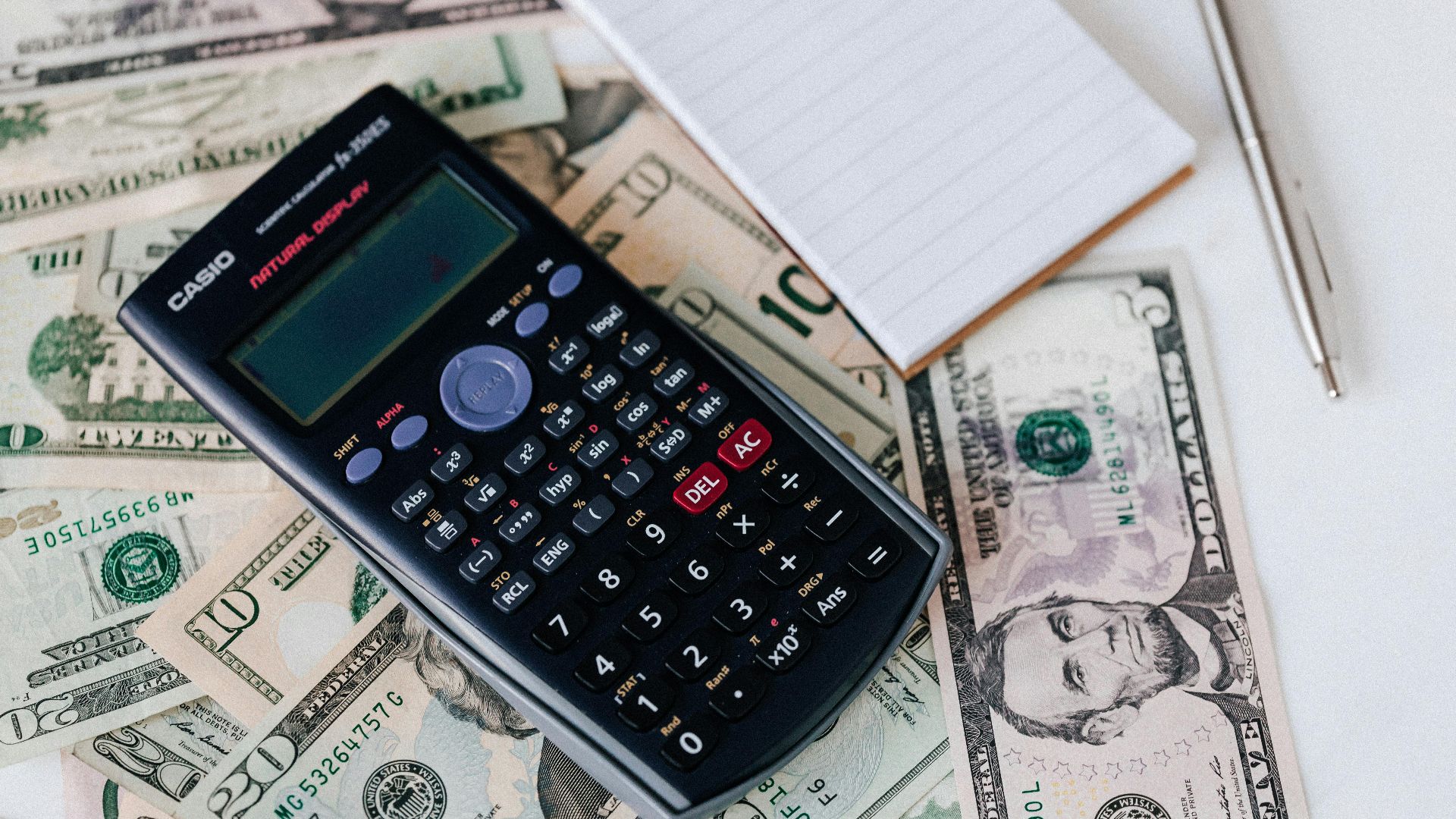 From above electronic calculator and notepad placed over United States dollar bills together with metallic pen for budget planning and calculation