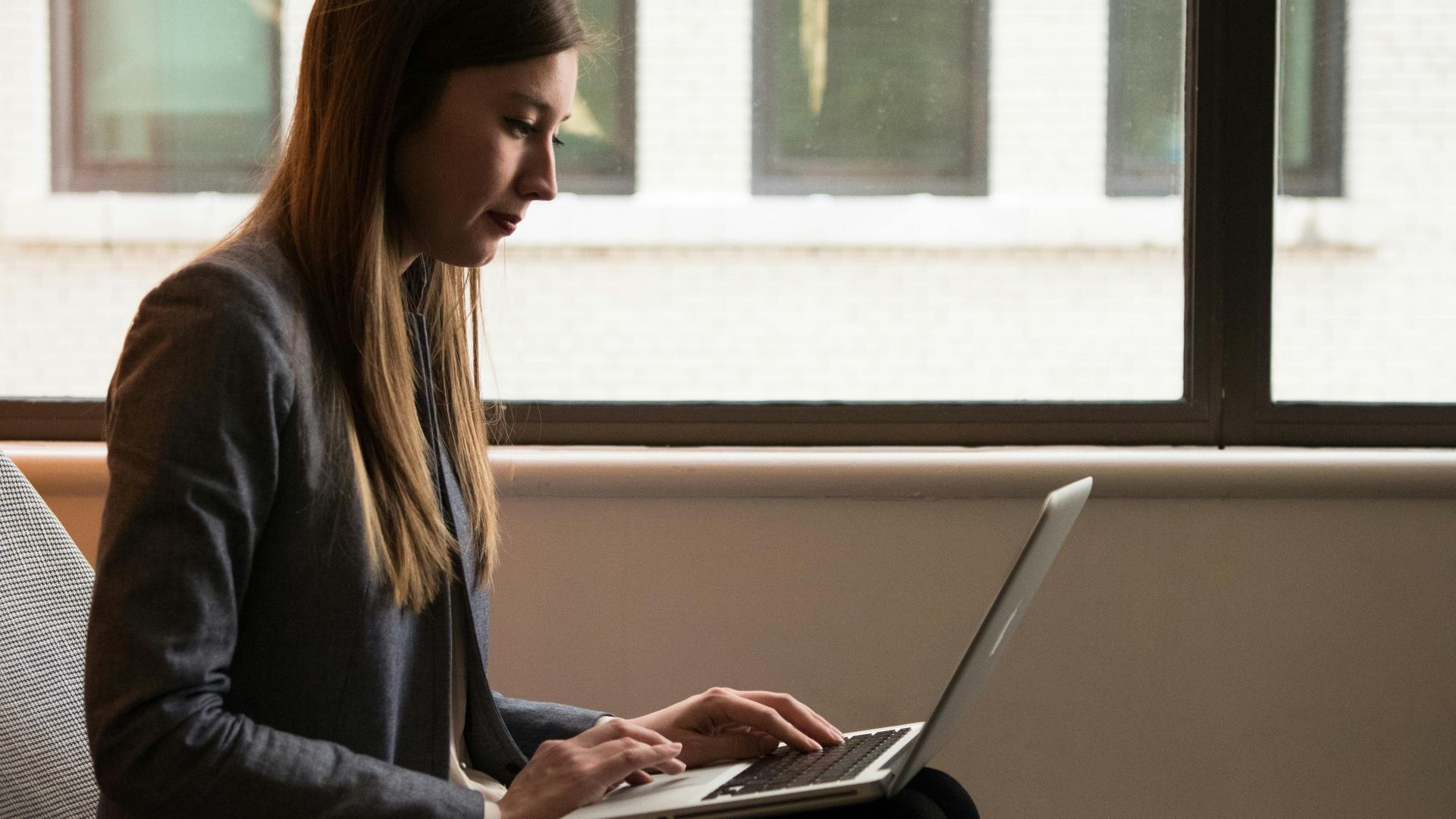 Woman sitting on a chair using a laptop by the window, engaged in remote work.