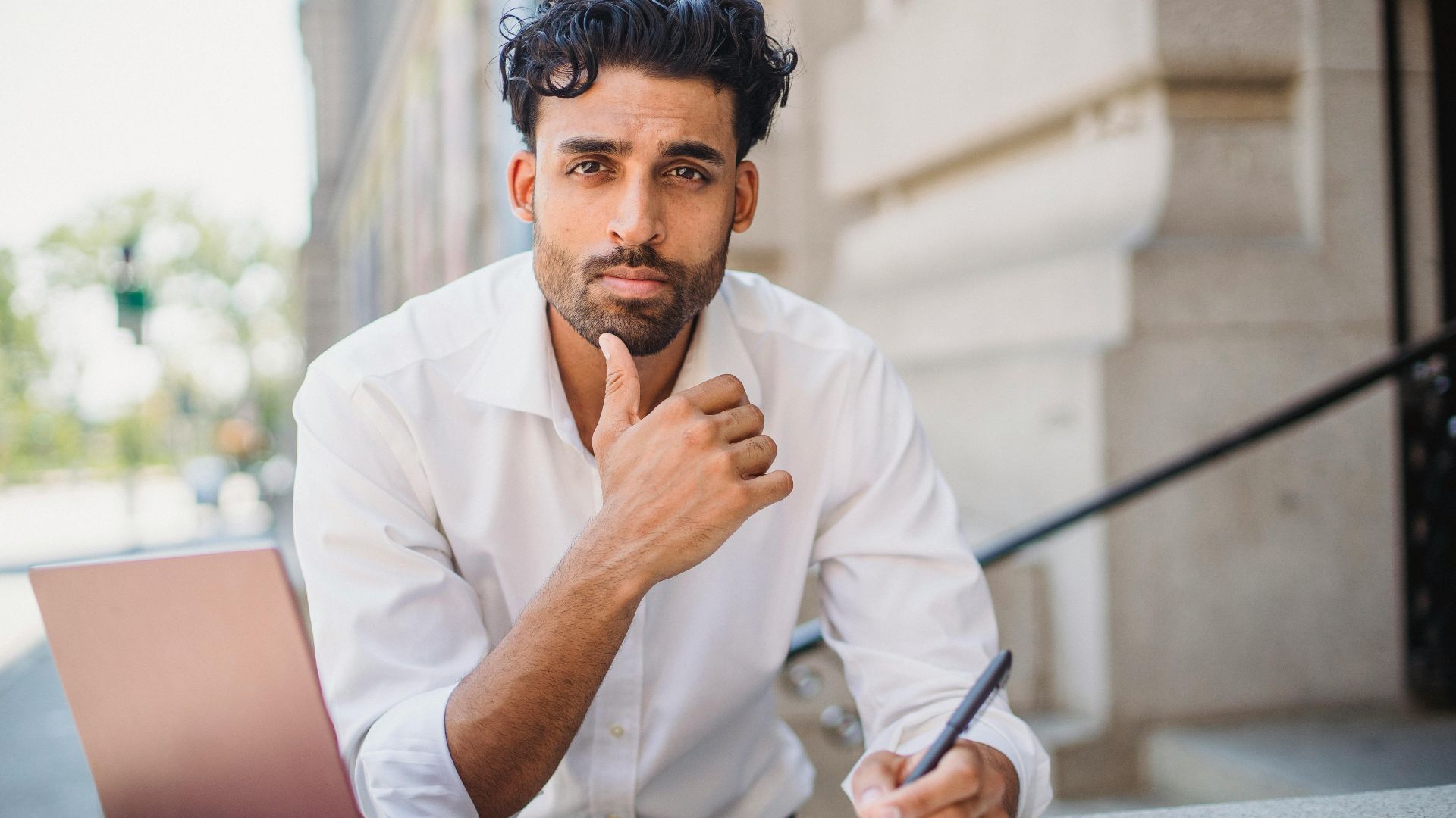 A man in formalwear thinking while writing in a notebook with a laptop outdoors.