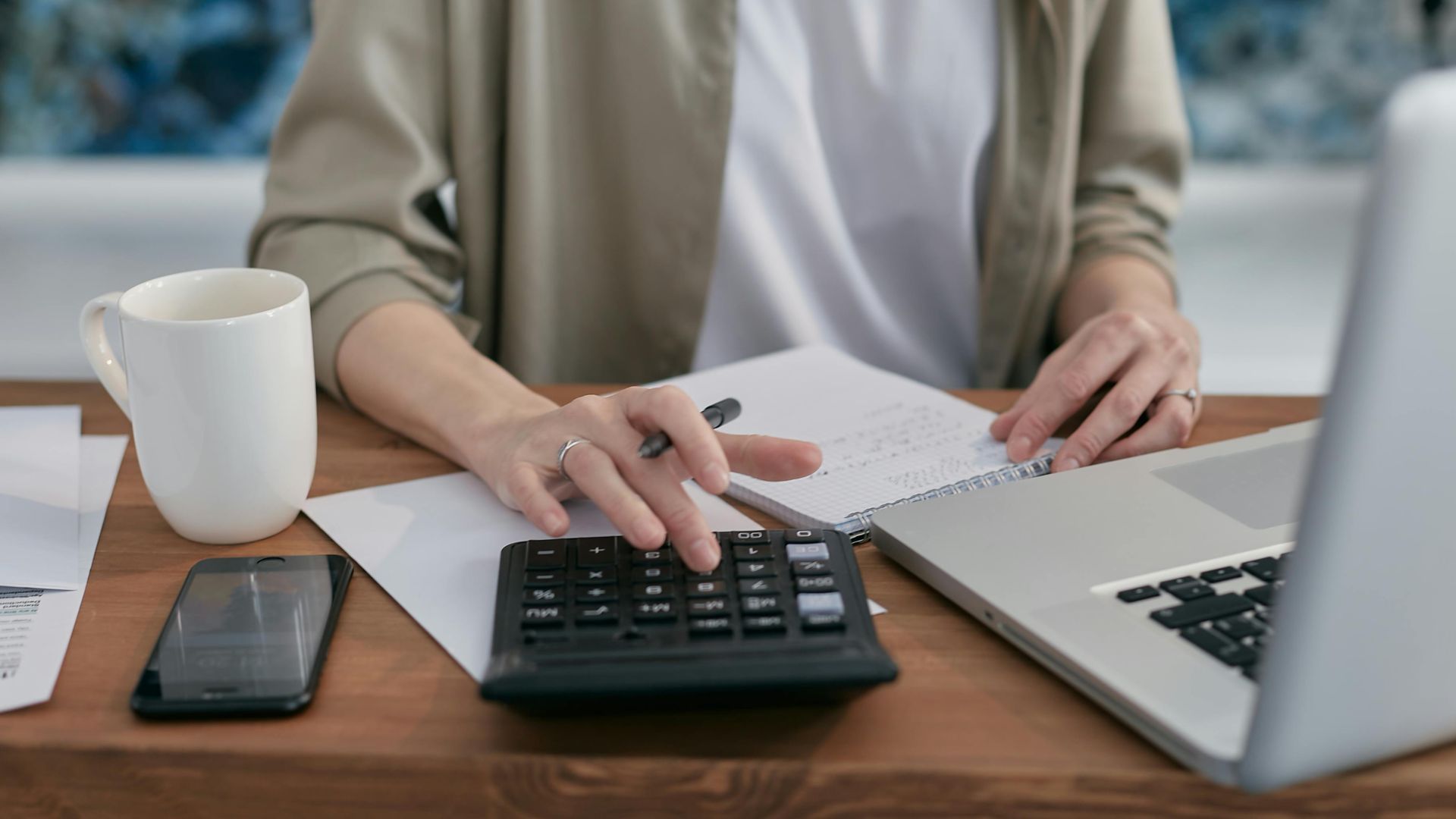 A person using a calculator and laptop while taking notes at a wooden desk.
