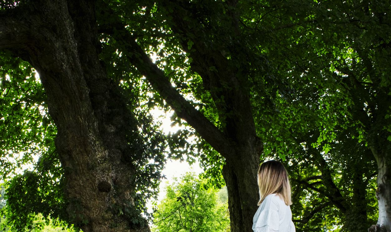 woman in white long sleeve dress standing on green grass field near brown tree during daytime