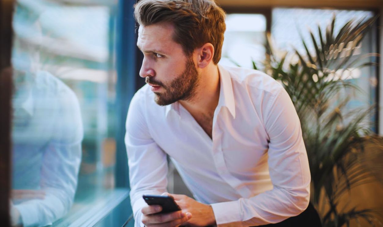 A Man Leaning on Glass Window Holding a Smartphone