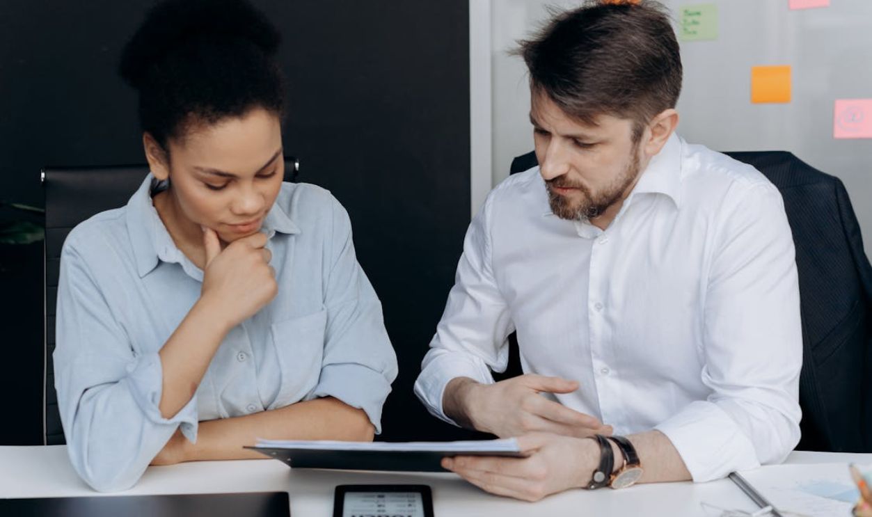 A Man and a Woman Looking at the Document on the Clipboard while Sitting Next to Each Other