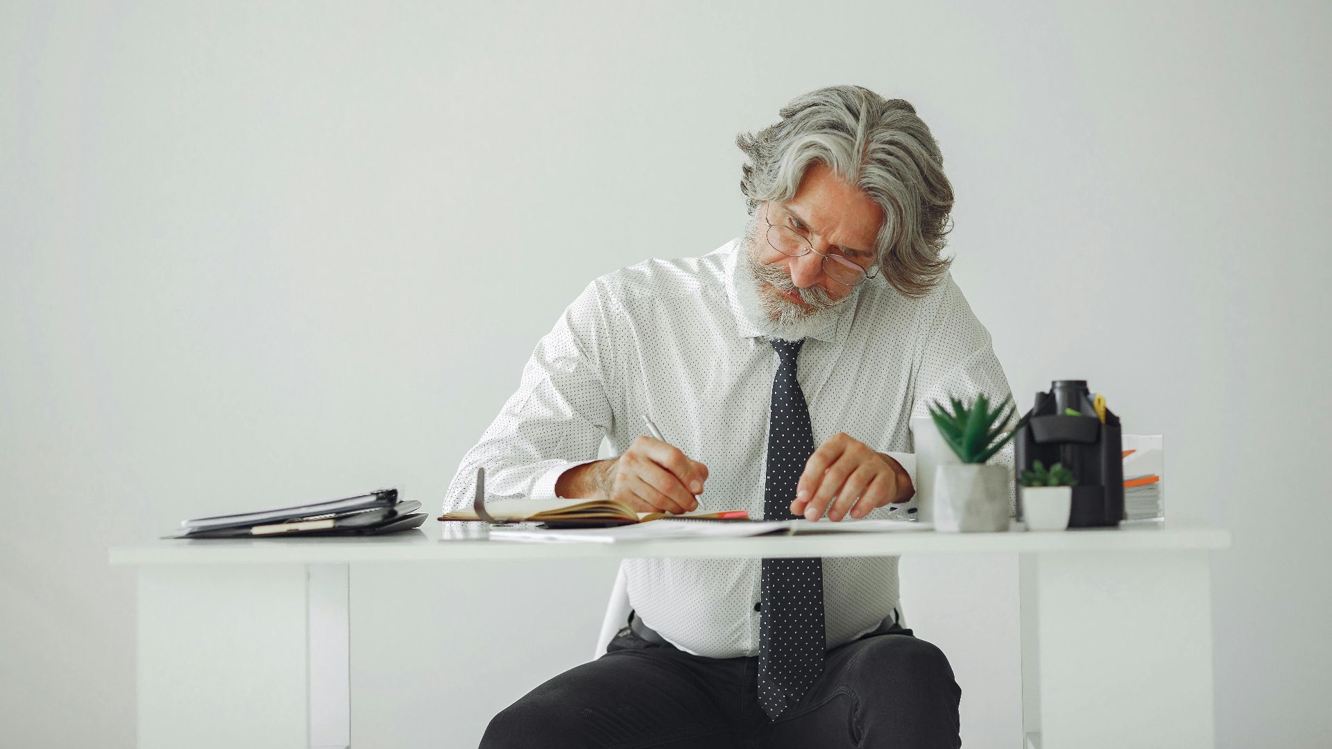 Elderly businessman with gray hair writes at a desk in a modern office.