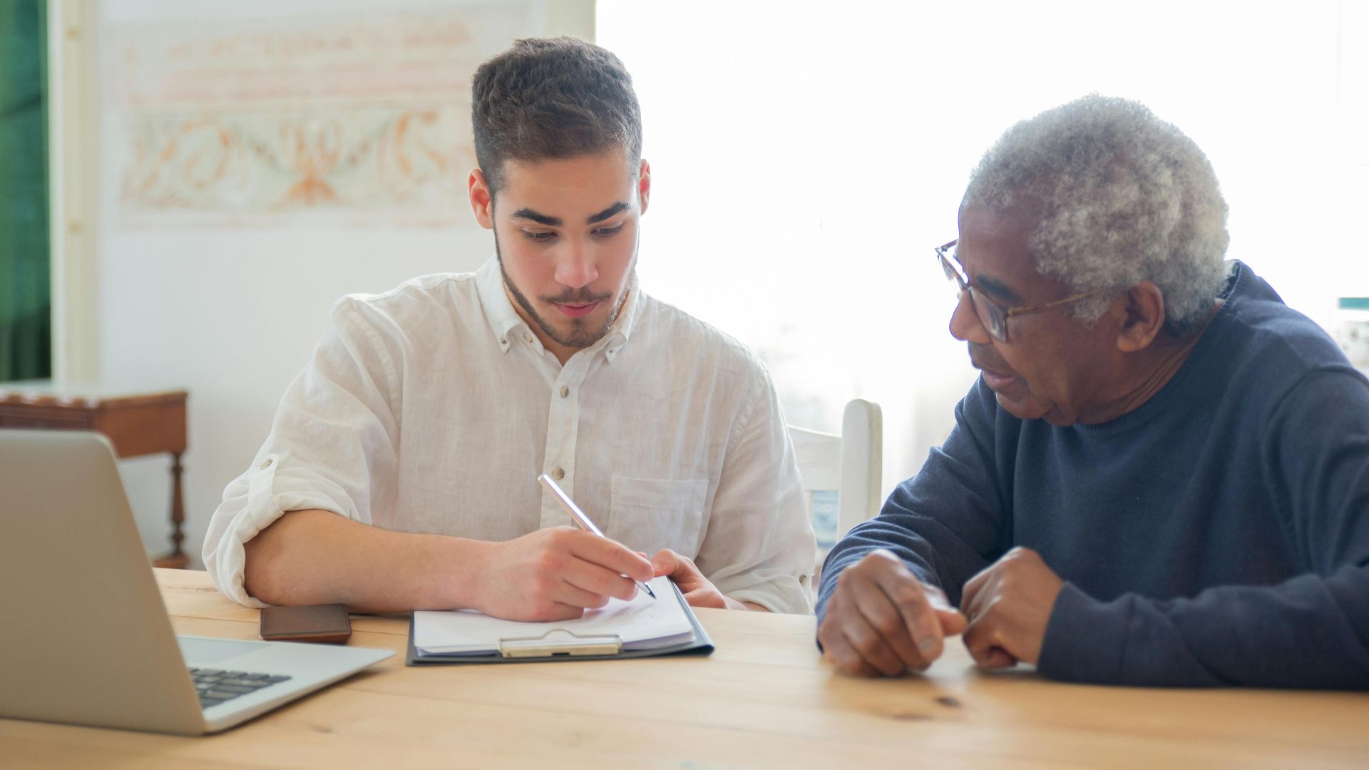 Young man helps elderly man with documents at a desk indoors, promoting collaboration.