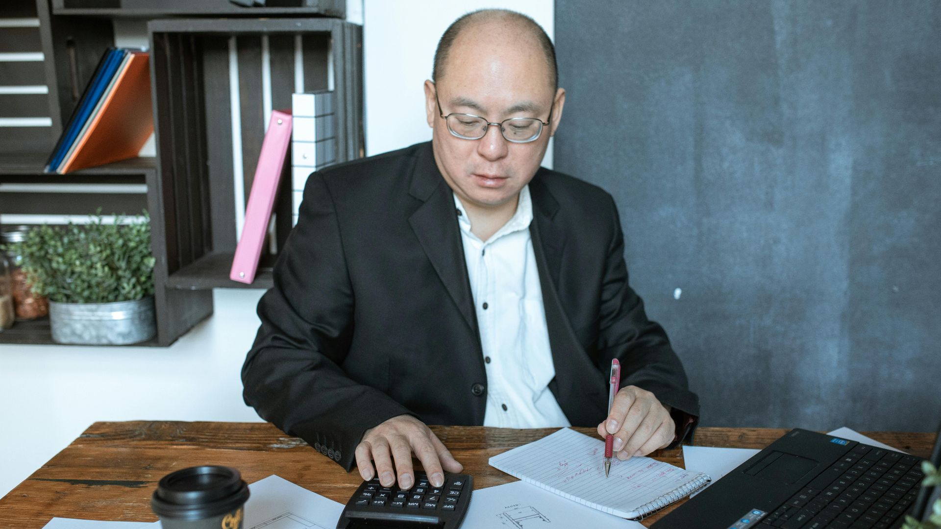 Asian businessman working with calculator and notes at his desk in a modern office space.