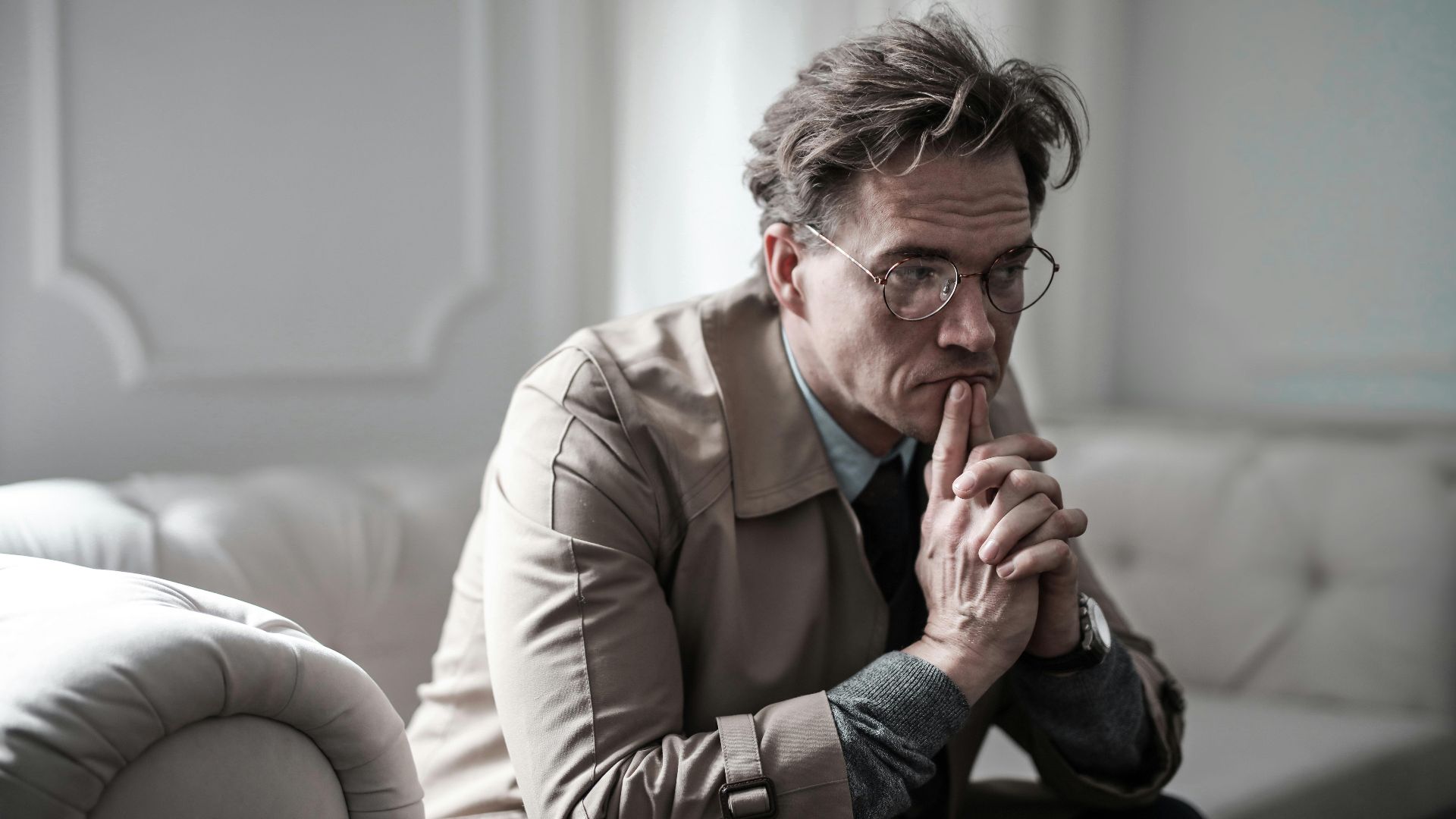 A thoughtful man wearing glasses sits on a sofa in a classic interior, lost in deep contemplation.