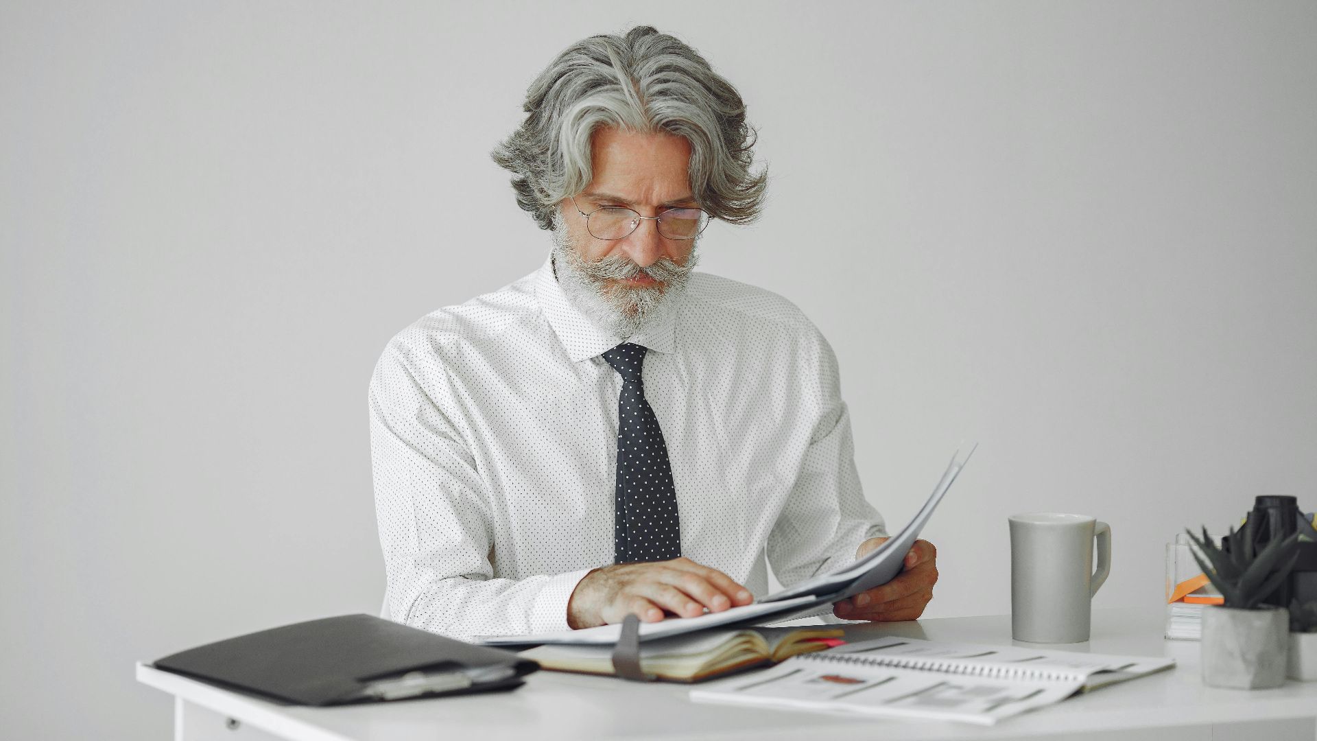An elderly businessman reviewing documents at his desk in a modern office setting.