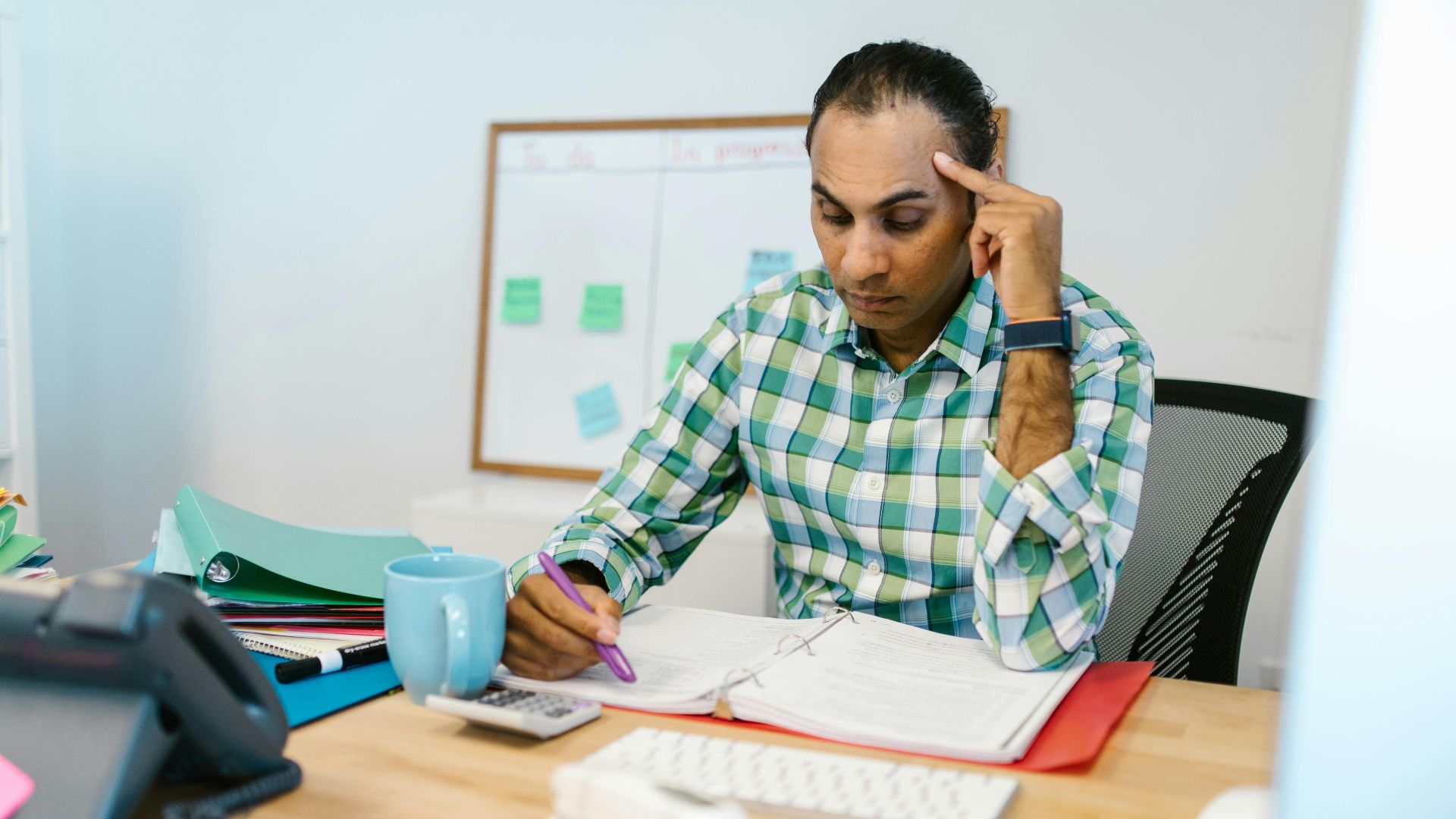A man sits at an office desk deeply engaged in reviewing papers, highlighting with determination.