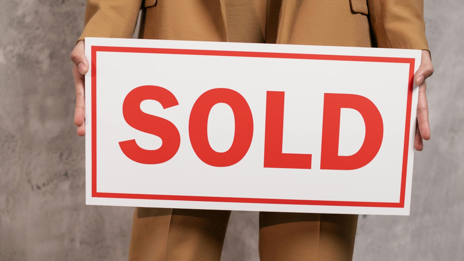 Close-up of real estate agent with sold sign in brown suit indoors.