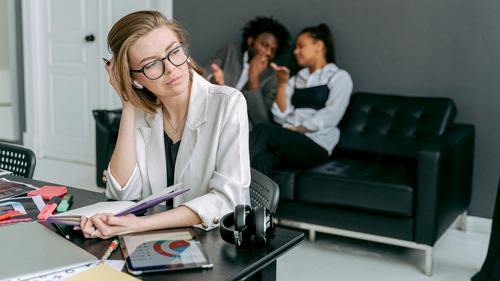 A woman appears stressed at her desk, with colleagues gossiping in the background.