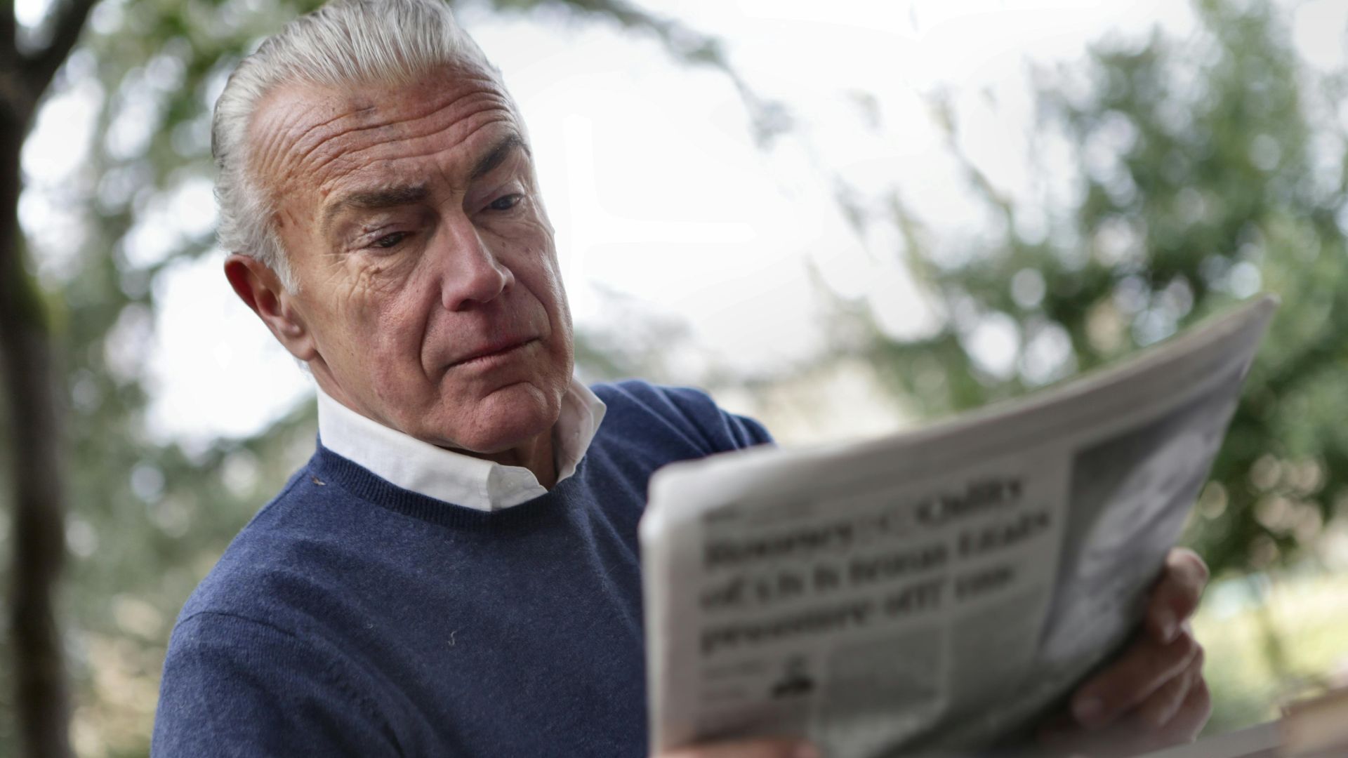 Elderly man reading a newspaper outdoors, wearing a blue sweater, showing serious expression.