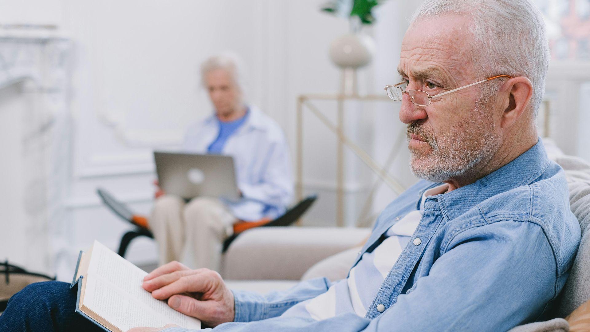 Senior man reading a book while an elderly woman uses a laptop in a cozy indoor setting.