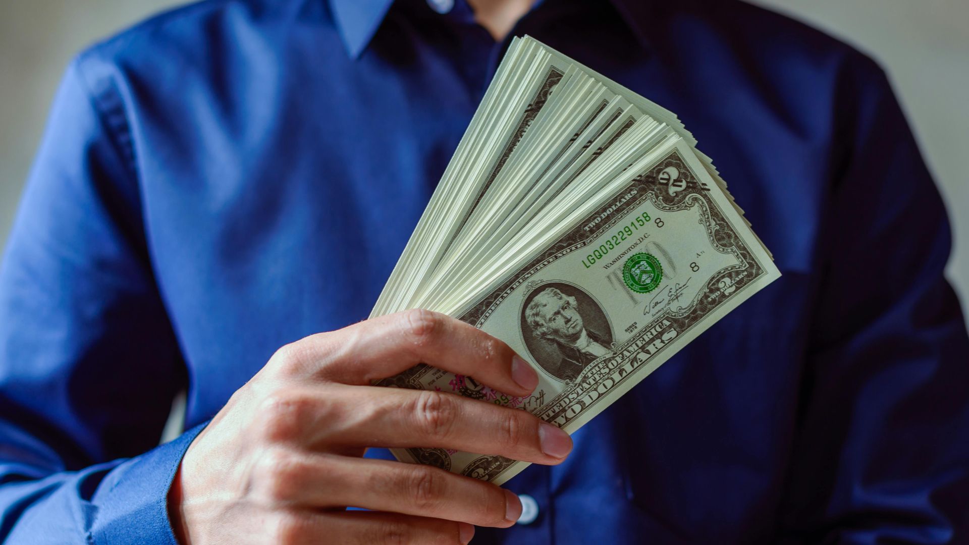 Close-up of a person in blue shirt holding multiple US dollar bills.
