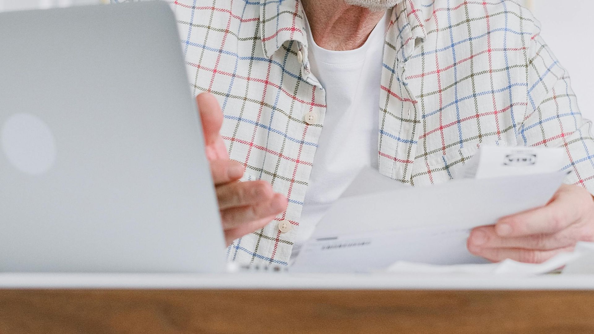 Elderly man reading papers at home with a laptop, emphasizing financial planning.