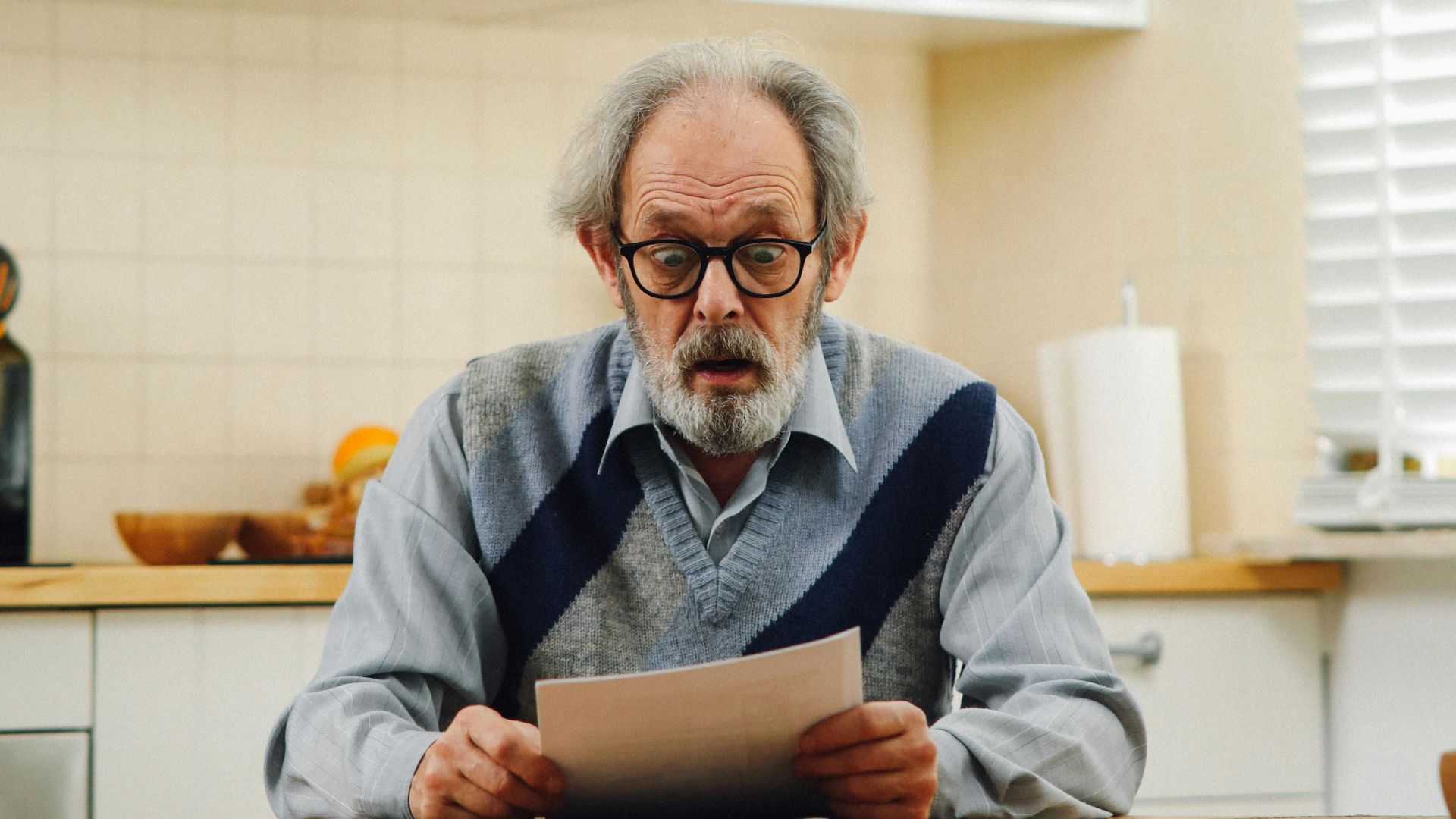 Senior man with eyeglasses reading a document at kitchen table, reflecting surprise.