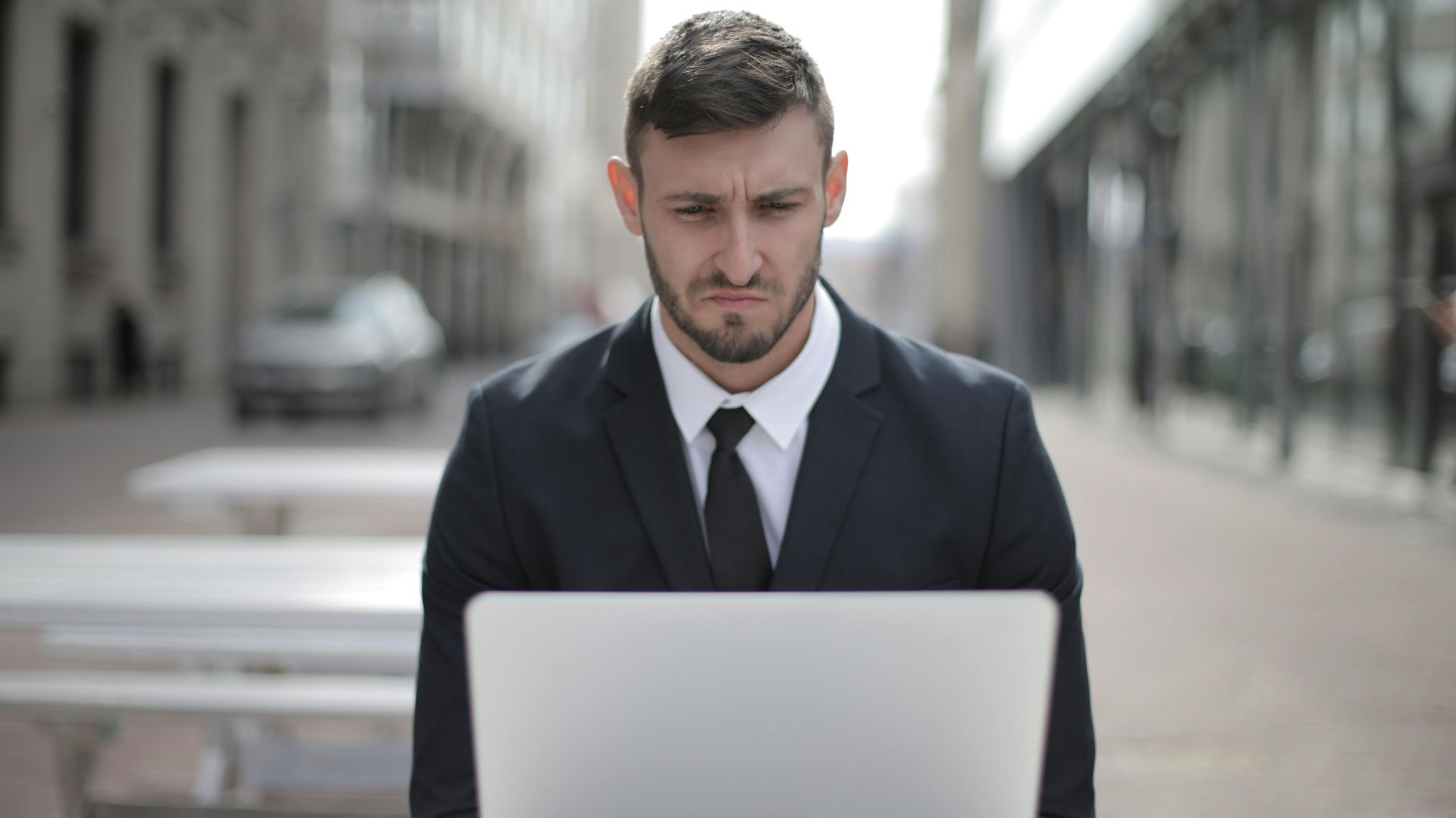 A focused businessman in a suit works on his laptop outdoors in an urban setting.