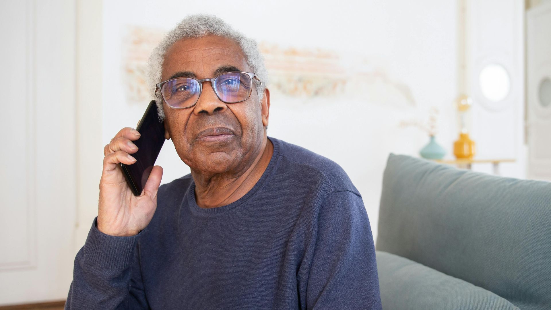 Senior man wearing glasses making a phone call while sitting on a sofa at home.