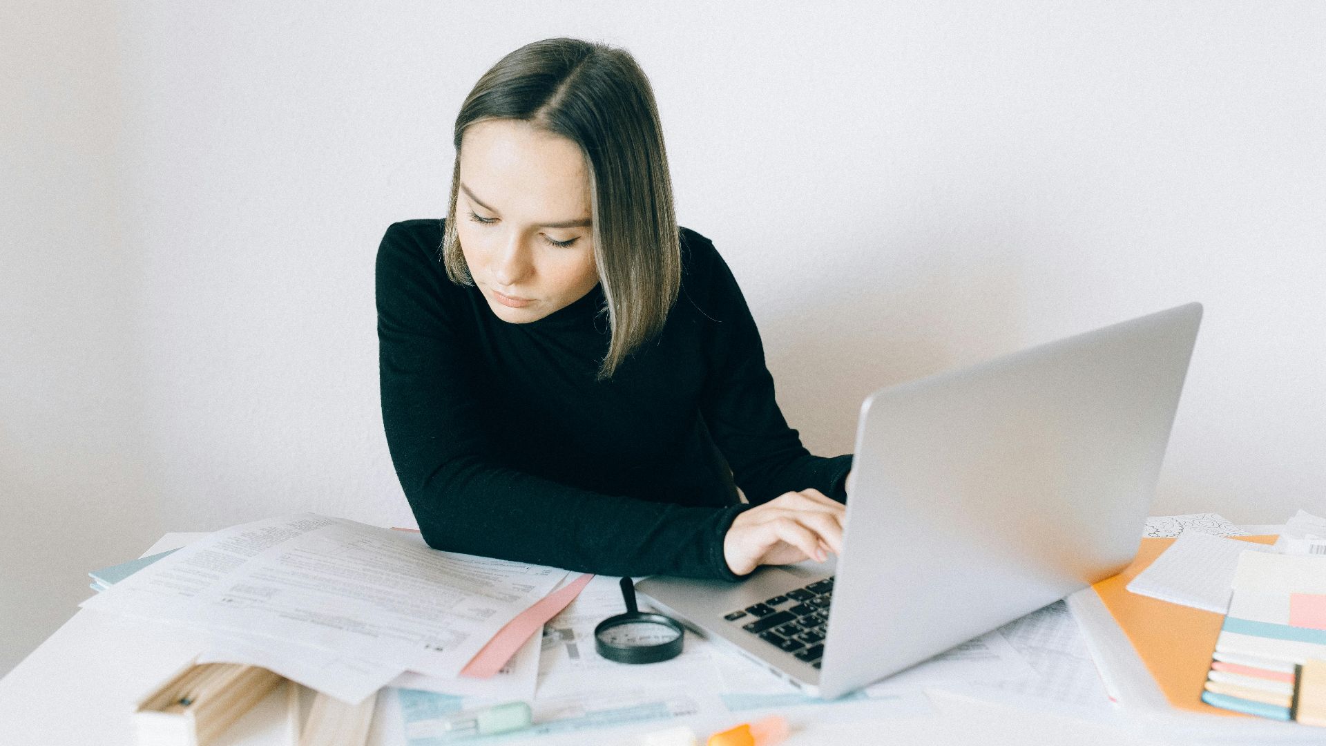 Focused woman researching on a laptop, surrounded by paperwork in a modern office.