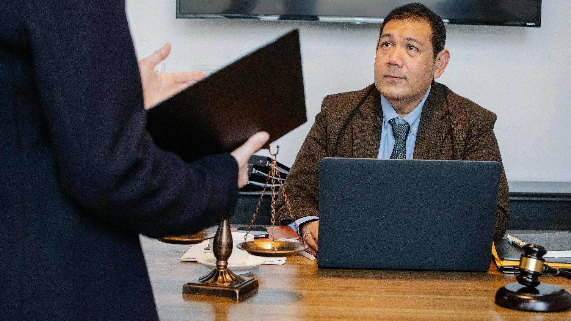 Crop unrecognizable employee representing new case details to concentrated middle aged ethnic lawyer sitting at table with laptop gavel and justice scales