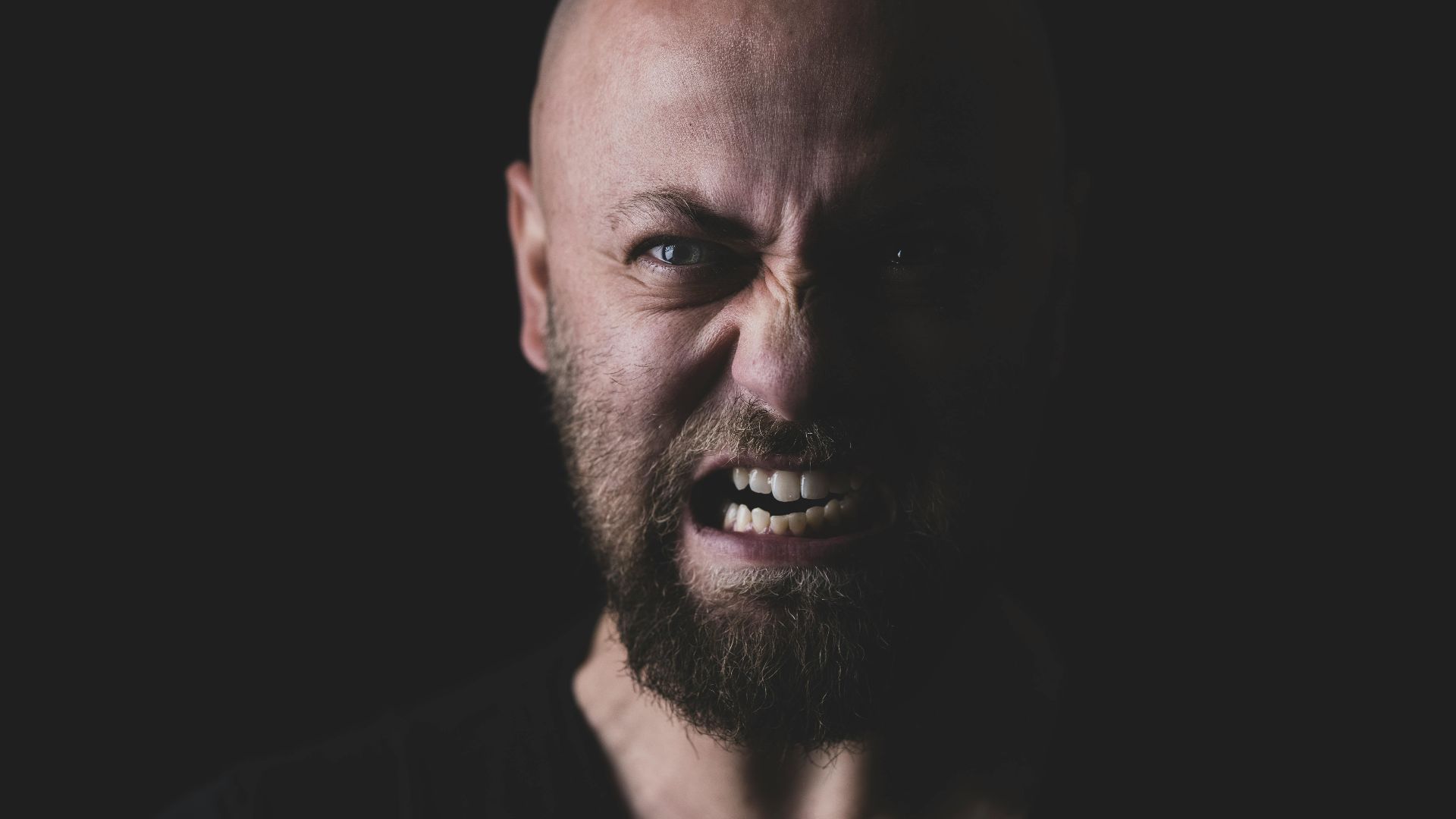 Close-up of a bald and bearded man showing a fierce expression in a dark setting.